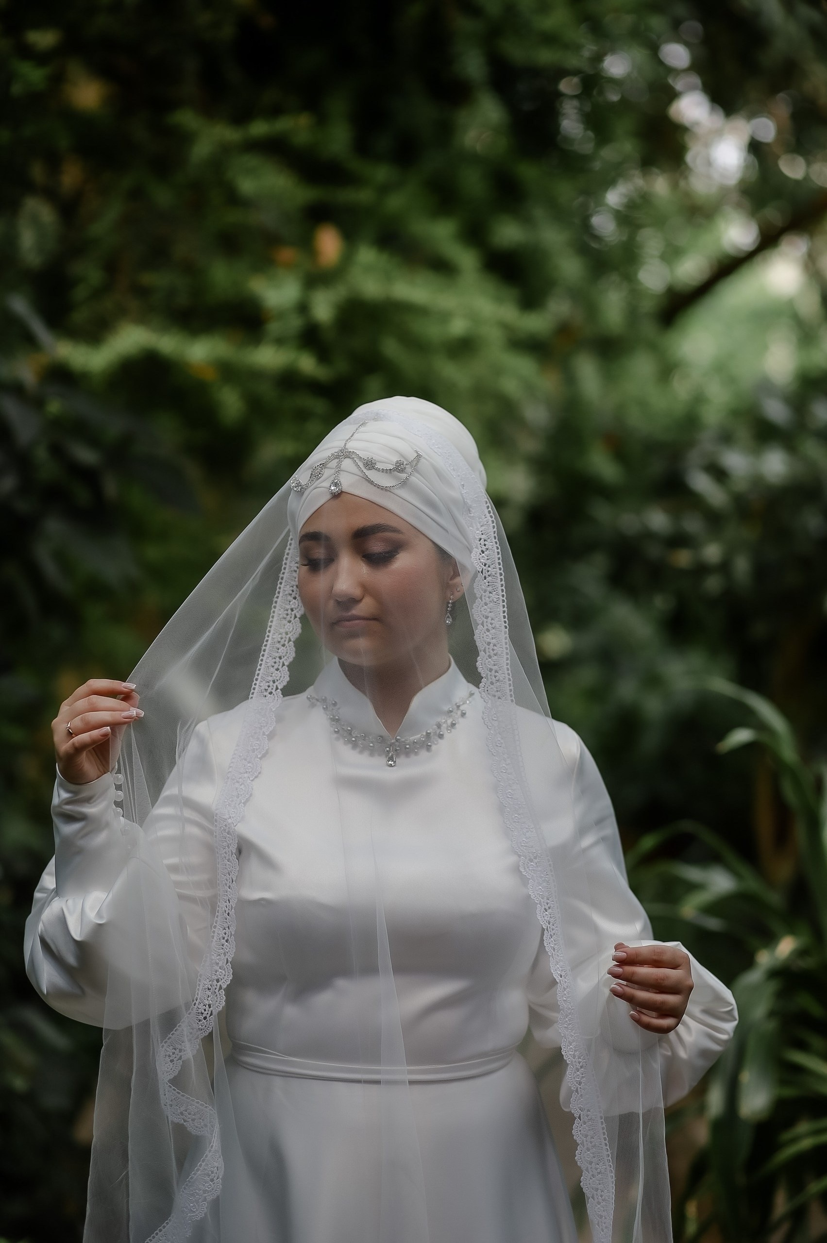Невеста в белом платье и платке с вуалью. Стоит среди зелени, выглядит задумчивой.