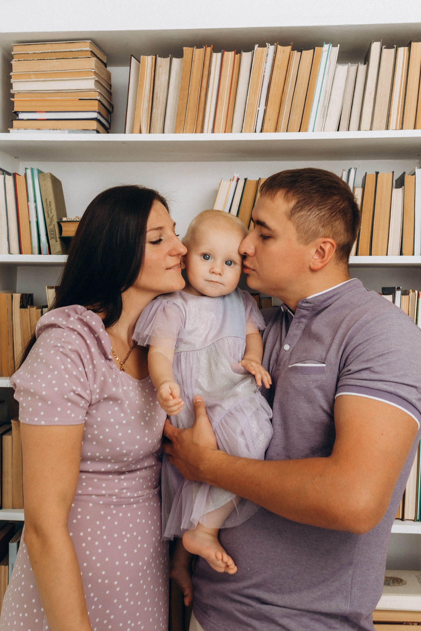 Родители целуют годовалую дочку на фоне книжных полок семейная фотосессия в Новосибирске