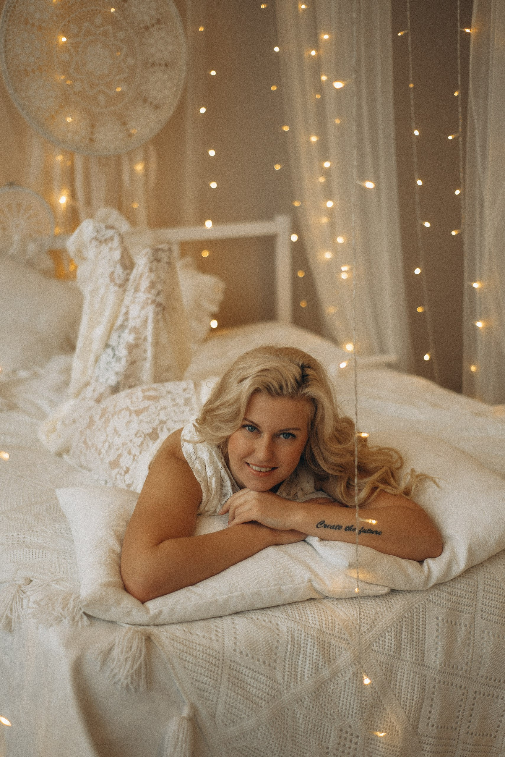 Улыбающийся портрет Натали Каролин на кровати. Модель лежит на животе, скрестив руки на подушке, игривый бохо-образ. Мягкое освещение от огоньков на переднем плане. Женская фотосессия в Новосибирске