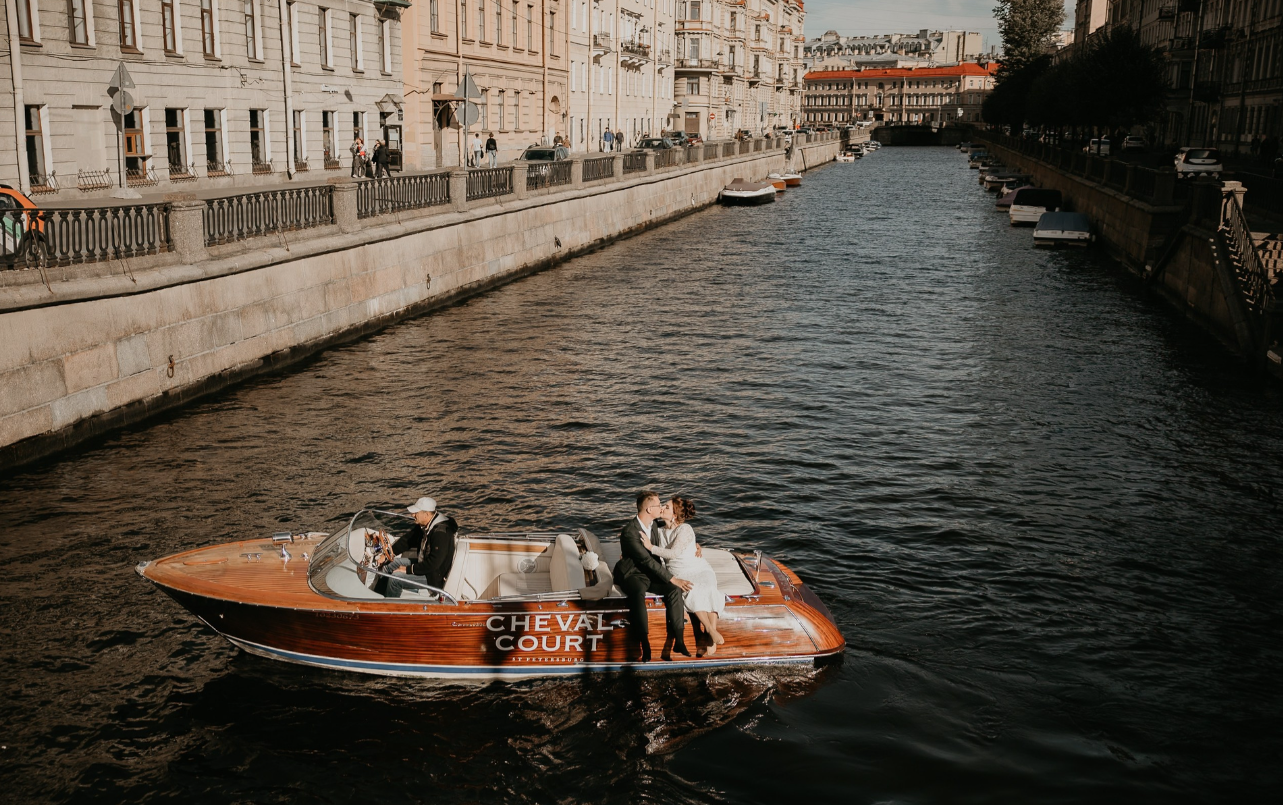 пара целуется на стильном катере, плывущем по каналу в городе. Атмосфера романтичная, вокруг старинные здания.