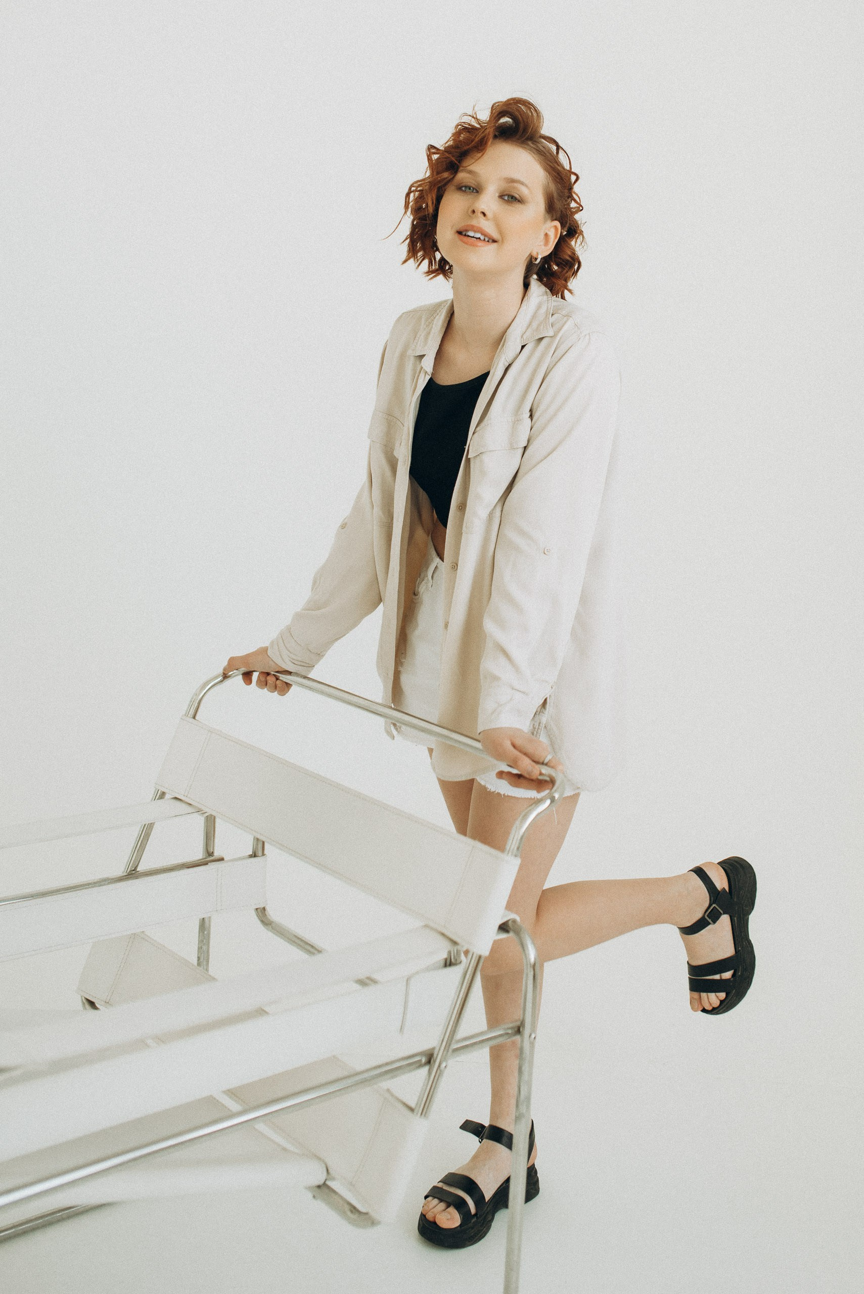 Эмоциональная поза модели у белого стула Баухаус. Модель в бежевой рубашке и шортах стоит с поднятой ногой. Весёлая и непринужденная фотосессия