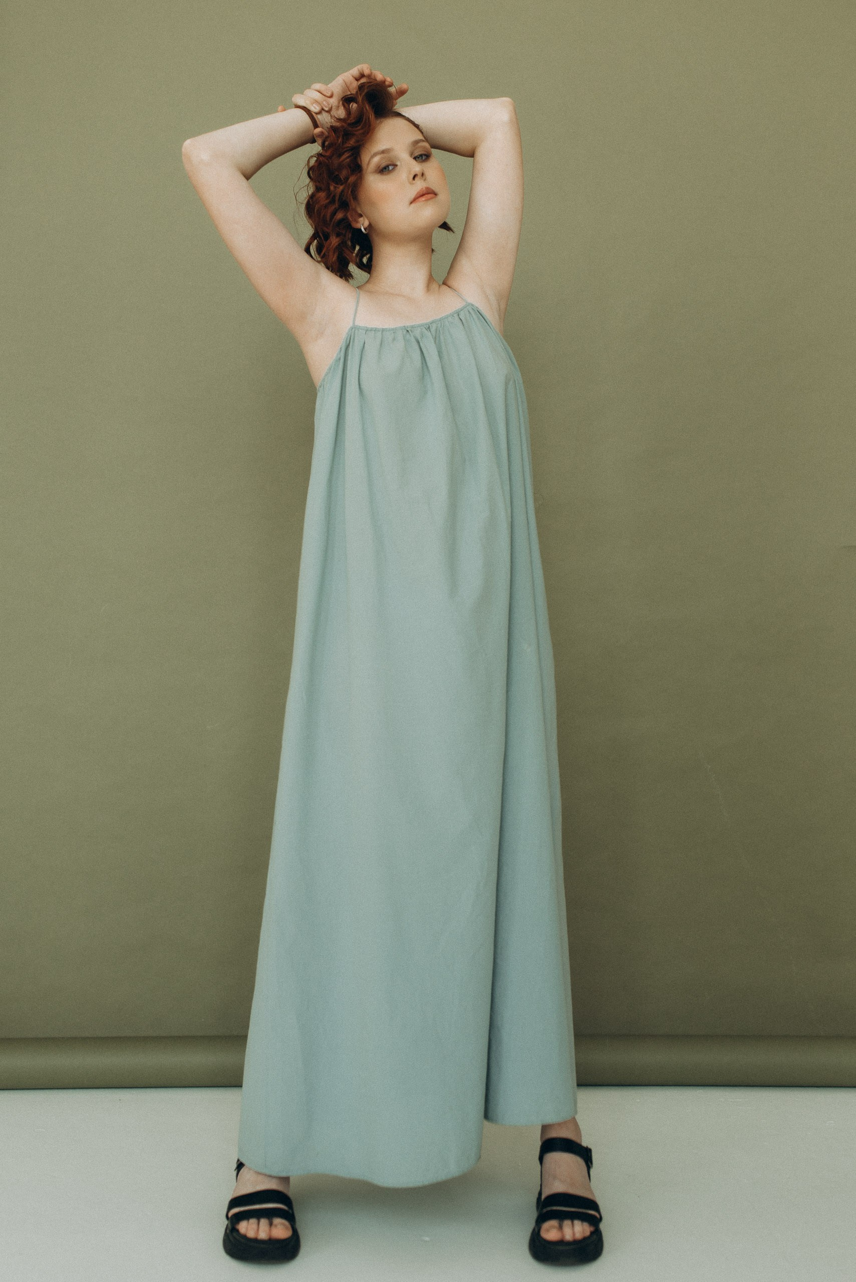 Стильная поза девушки в полный рост с руками за головой. Длинное платье и оливковый студийный фон. Пример качественного подросткового портрета
