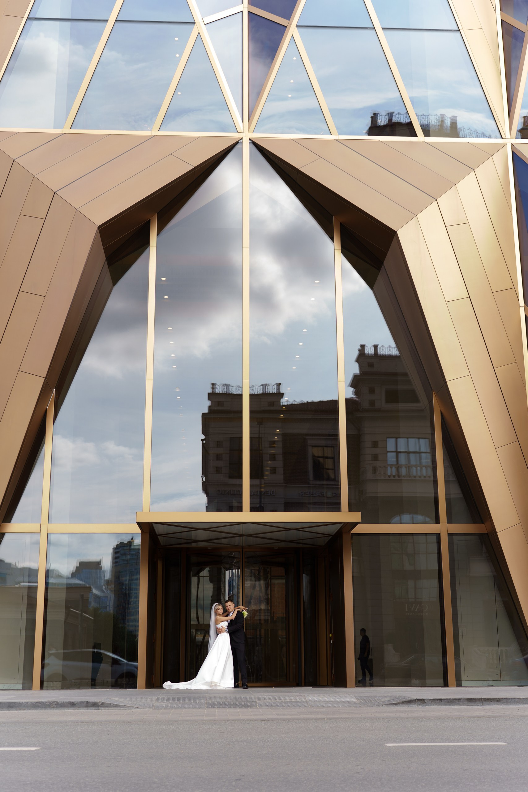 Жених и невеста обнимаются на фоне современного архитектурного шедевра — здания РМК в Екатеринбурге. Стеклянные панели фасада отражают небо и городские постройки, создавая динамичный фон для снимка | фотограф для семейного альбома Екатеринбург