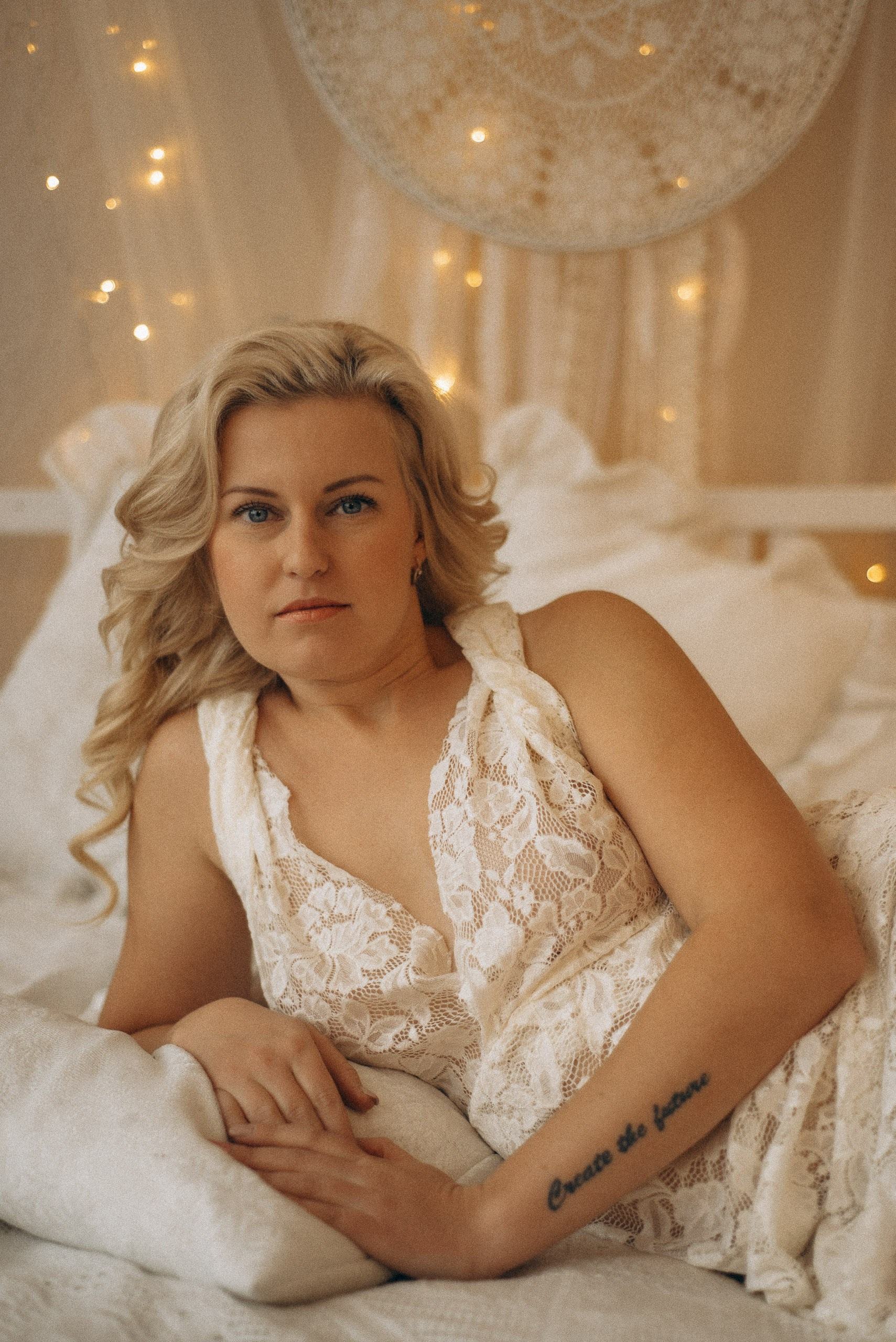 Крупный портрет Натали Каролин на кровати. Акцент на татуировке на руке и текстуре белого кружевного платья. Мягкий свет гирлянд на фоне бохо-студии. Женская фотосессия в Новосибирске