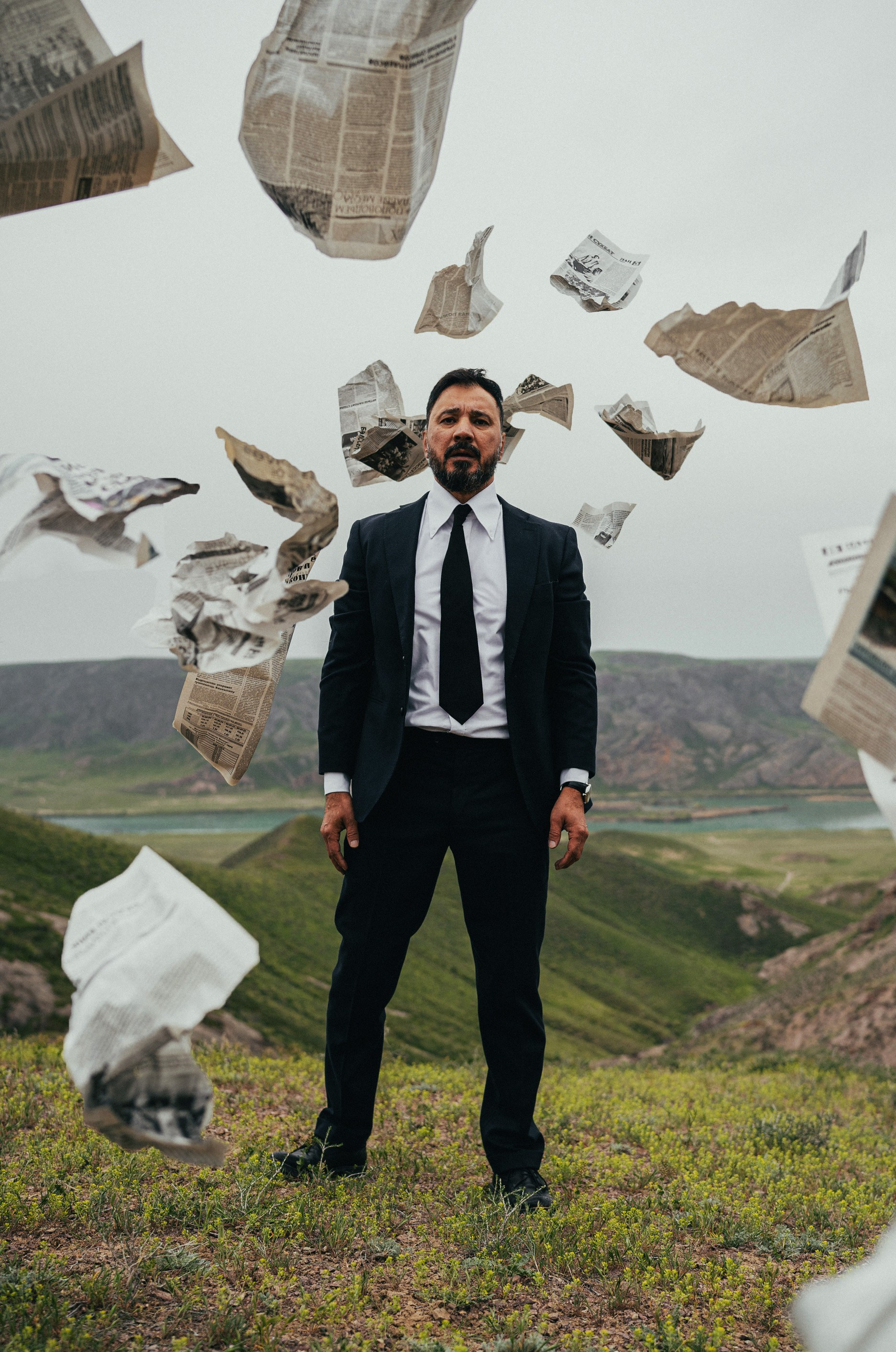 Мужчина в чёрном костюме стоит на холме, вокруг него в воздухе летают страницы газет