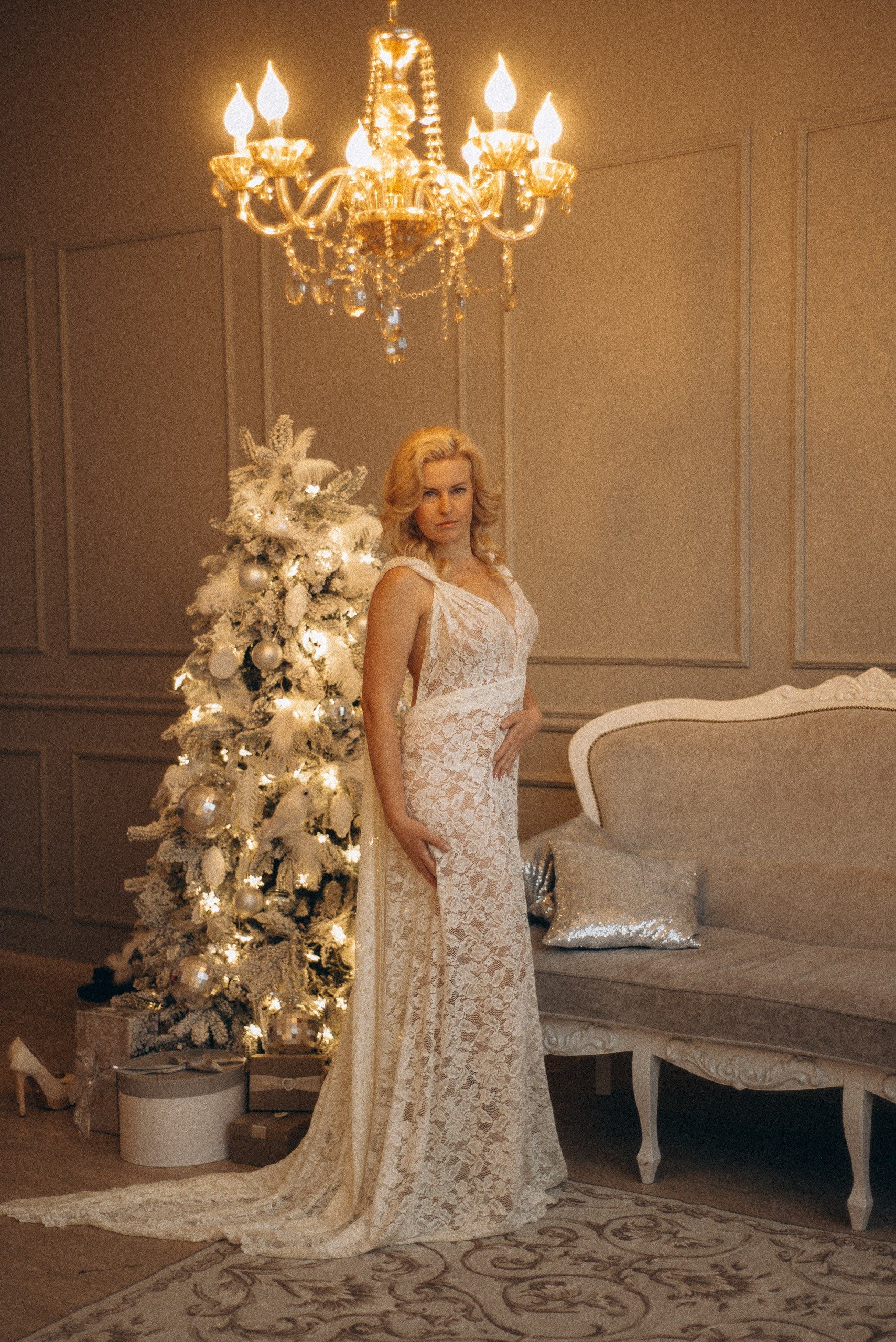 Натали Каролин в полный рост в роскошном классическом интерьере. Модель в белом кружевном платье со шлейфом стоит у украшенной новогодней елки. Классический диван и люстра. Праздничная женская фотосессия, фотограф Василий Дан