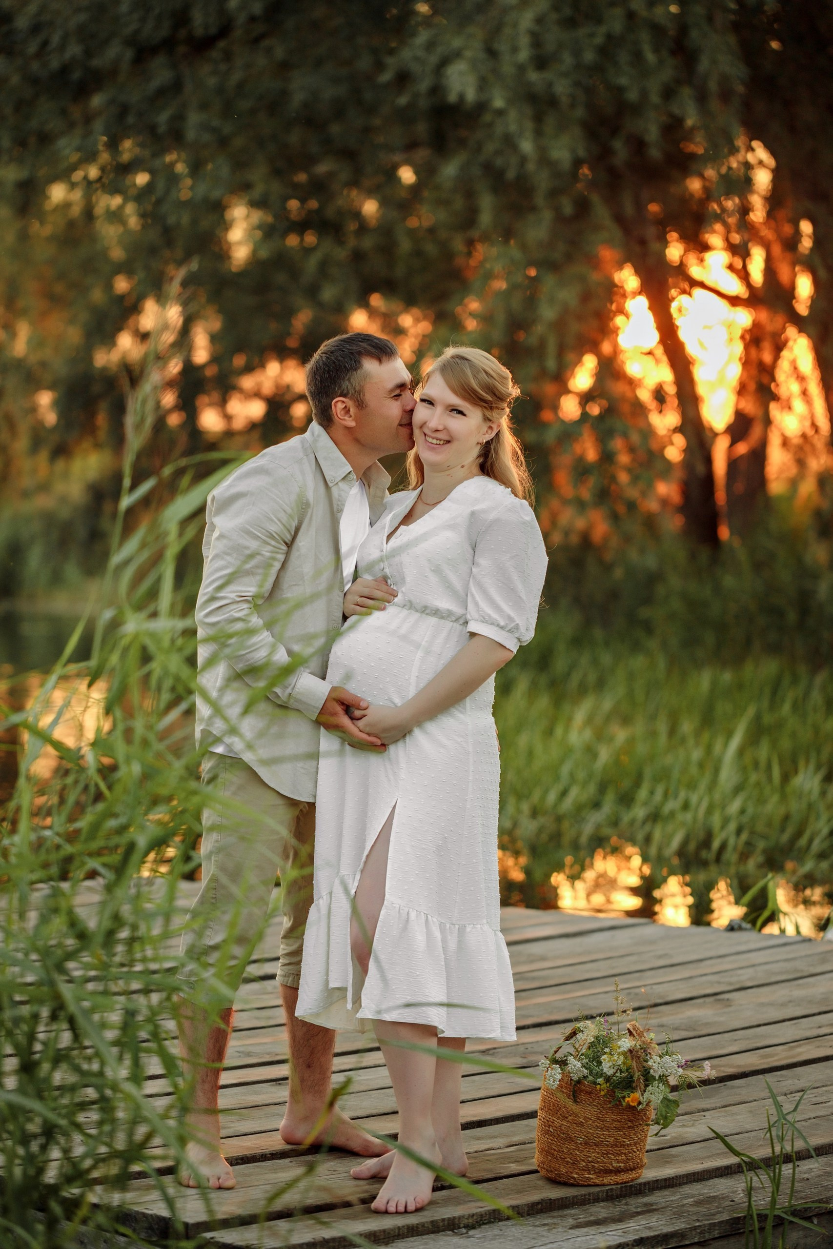 Беременная женщина и её муж на деревянном причале у воды. Мужчина целует жену в щёку, она улыбается. Закат, зелень вокруг.