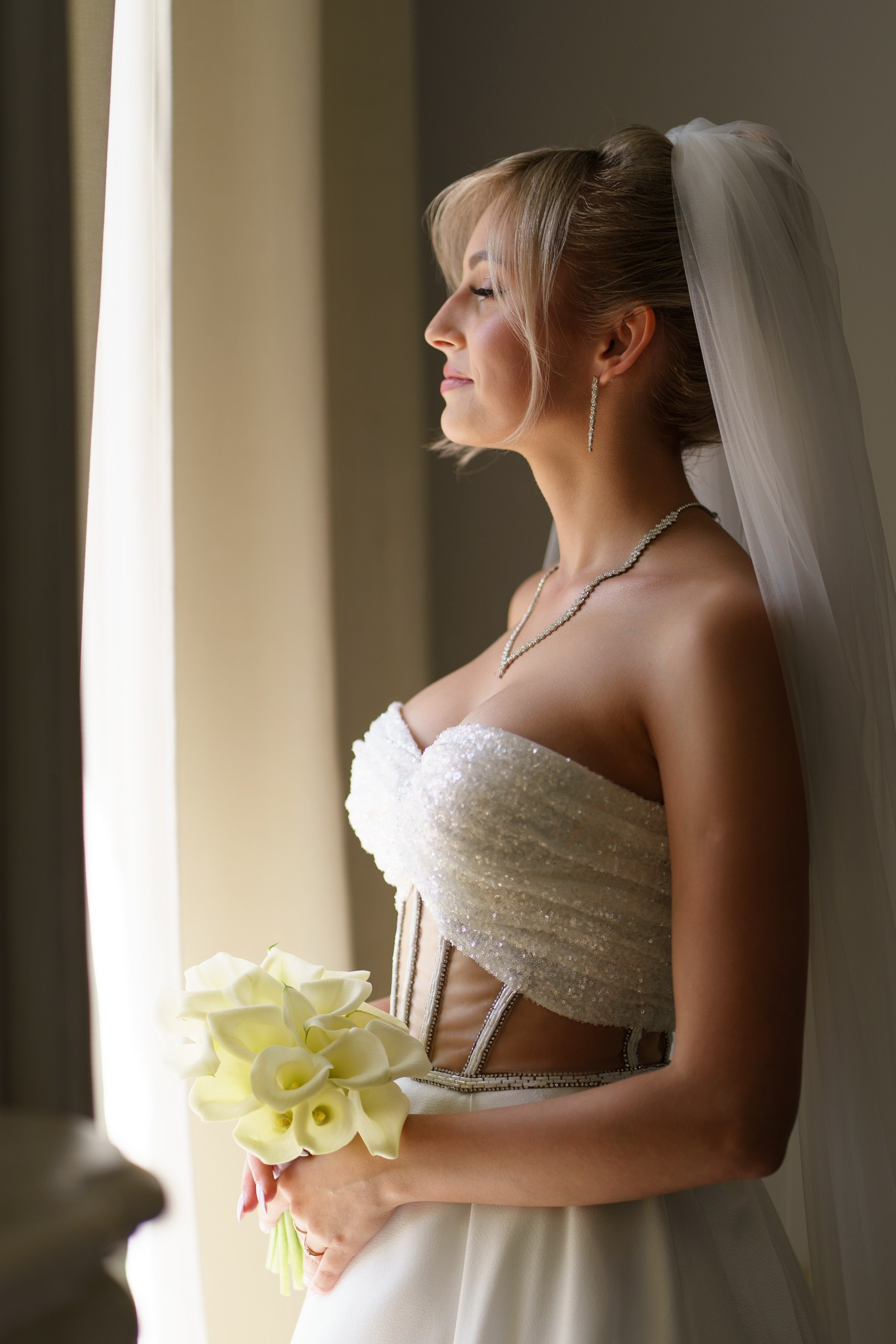Портрет невесты в день свадьбы: она одета в блестящее платье, дополненное фатой и украшениями. Букет белых калл завершает её образ. Снимок выполнен в профиль | портретная свадебная съемка Екатеринбург