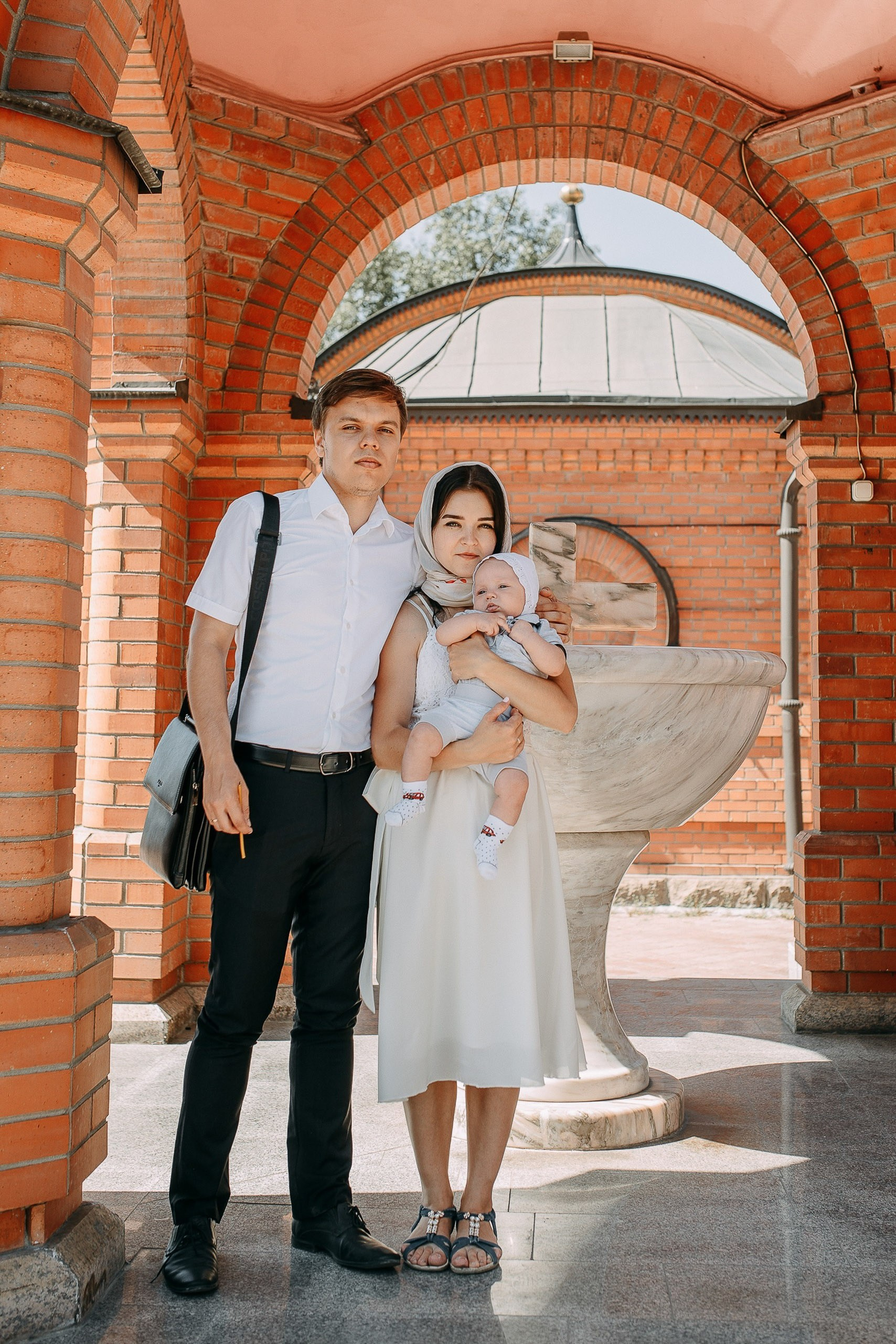 Семья из трех человек с младенцем на руках позирует под кирпичной аркой православного храма в солнечный день.