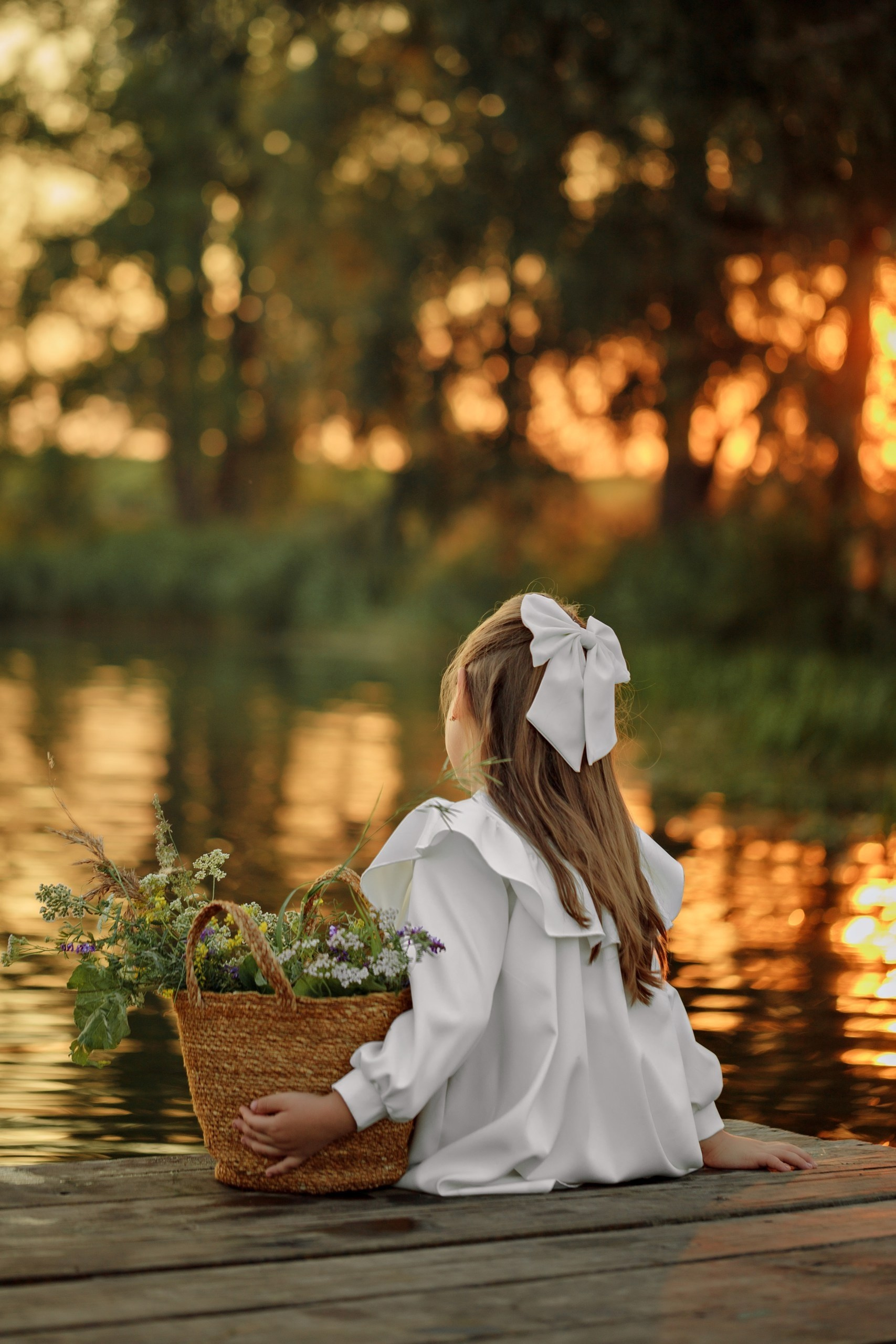 Девочка в белой блузке с бантом сидит на причале у реки. В руках плетёная корзина с полевыми цветами. Закат.