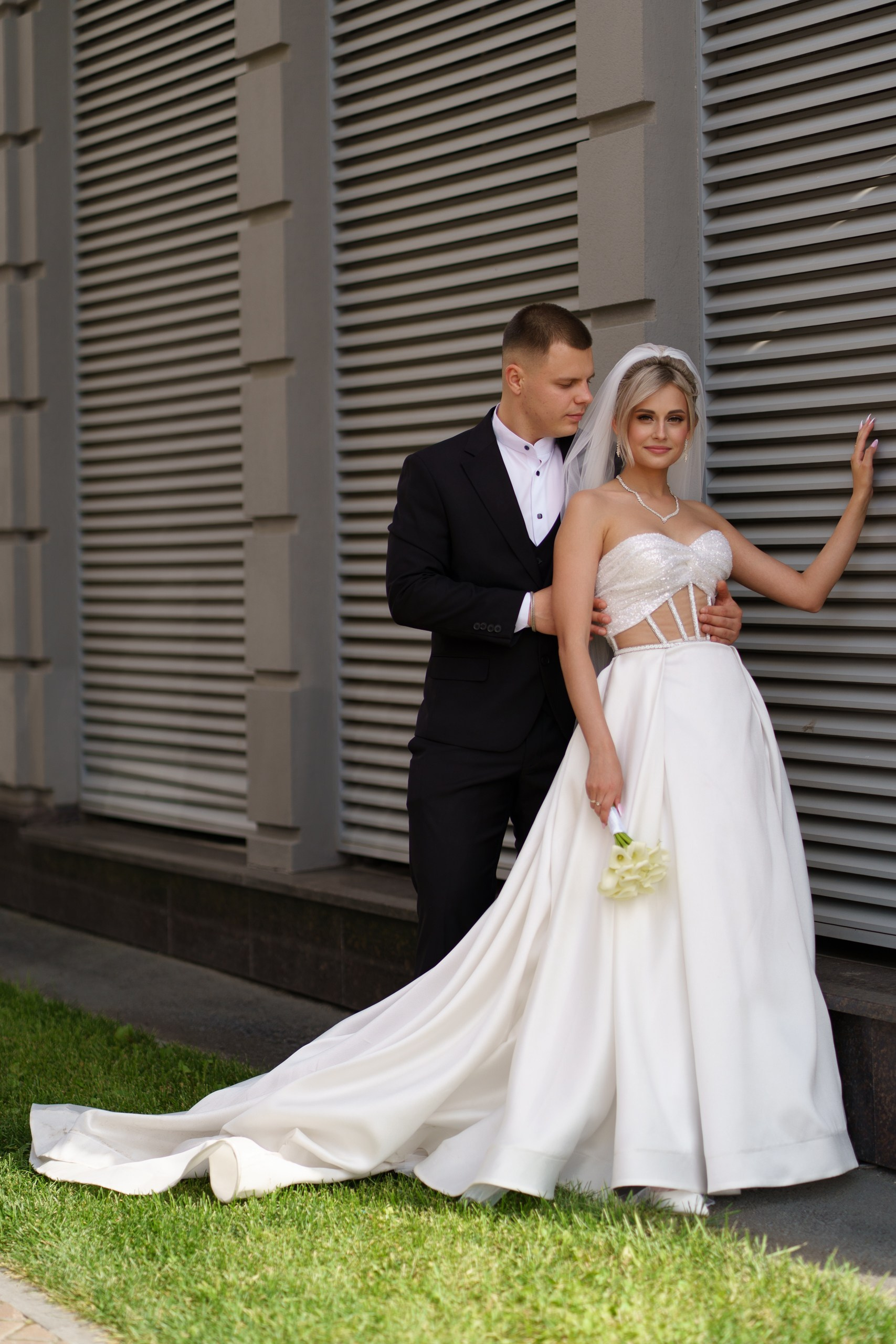 Невеста в роскошном свадебном платье с длинным шлейфом и фатой позирует, опираясь рукой о стену, рядом с ней — жених в классическом чёрном костюме | свадебный фотограф для торжеств Екатеринбург