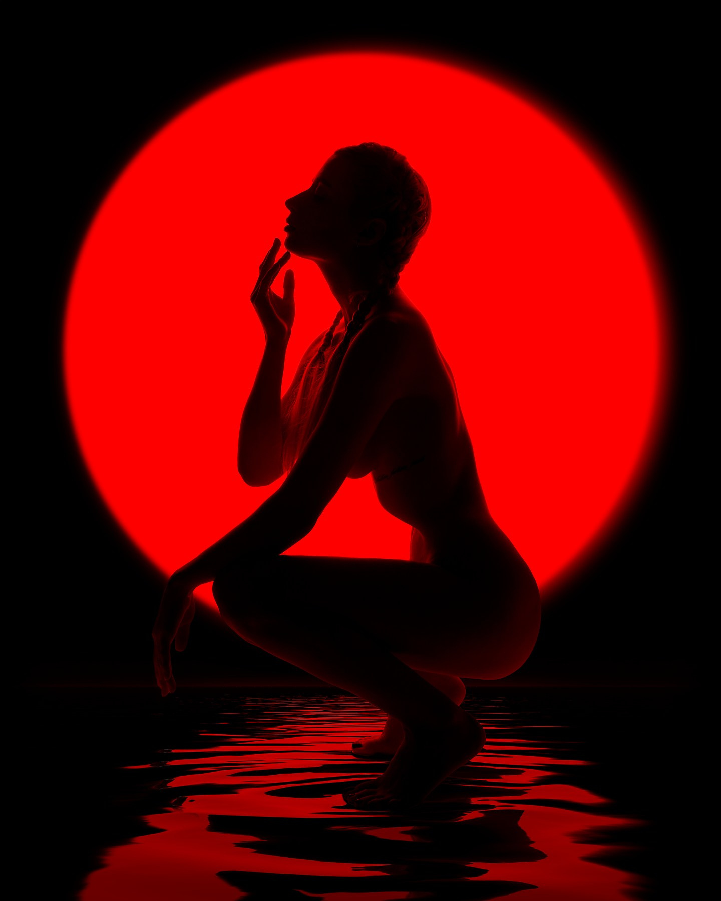 Творческая съёмка с элементами символизма: силуэт модели, сидящей на коленях, с поднятой рукой, на фоне яркого красного круга, напоминающего солнце. Модель окружена водной поверхностью, создающей отражения, что усиливает визуальный эффект. Сильный контраст света и тени подчёркивает драматичную атмосферу кадра