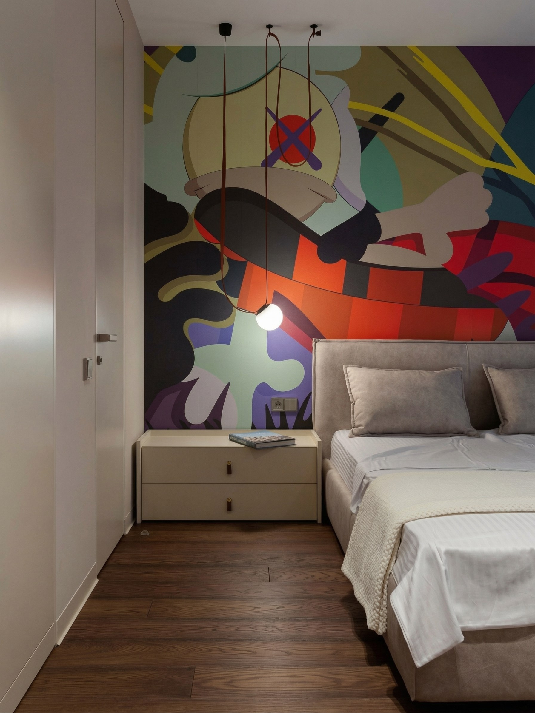 Современная спальня с артом KAWS от Валерии Котович: встроенный шкаф в потолок, дизайнерский подвесной светильник и темная паркетная доска.