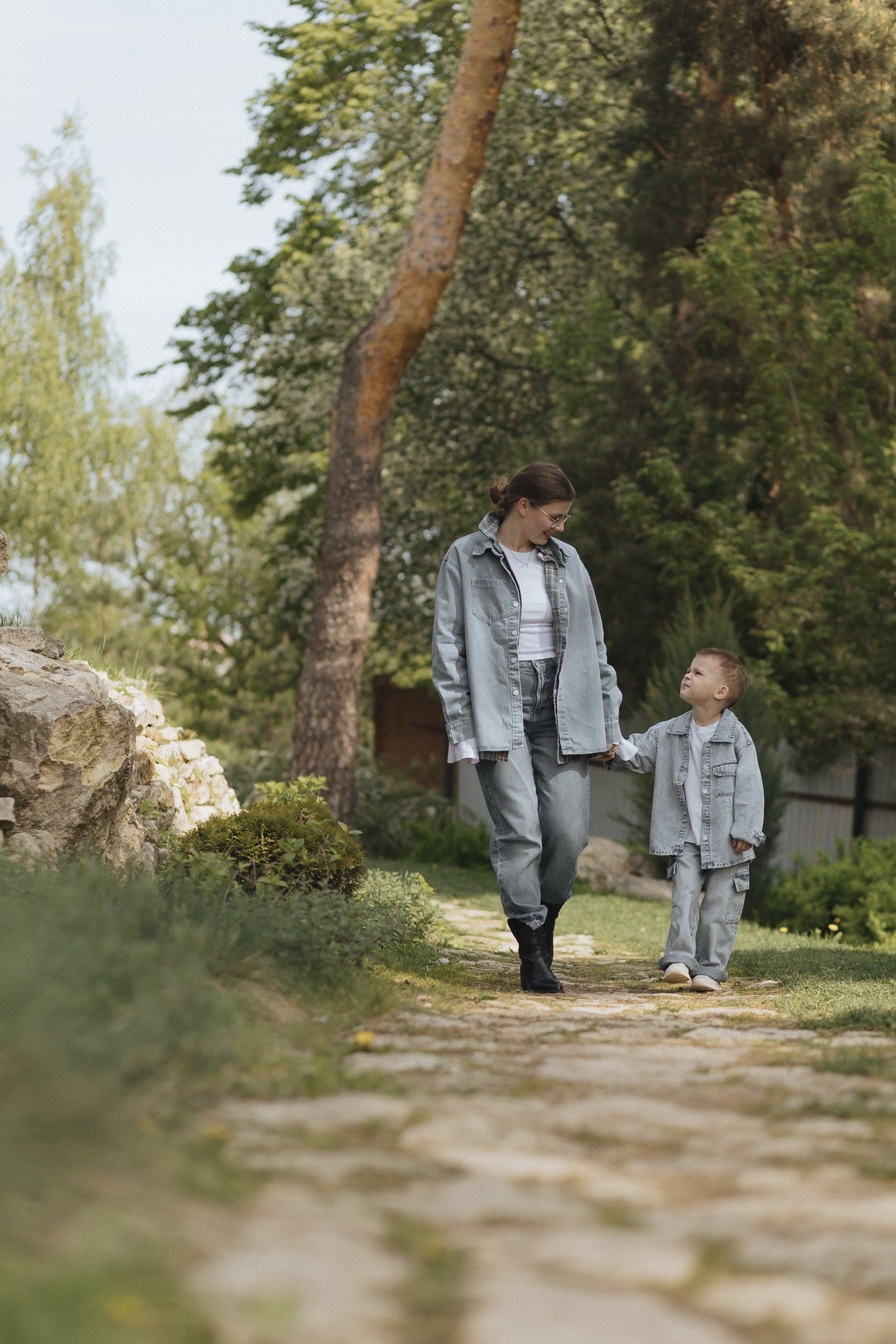Мама с малышом гуляют по садовой дорожке, держатся за руки. Вокруг — зелень, деревья.
