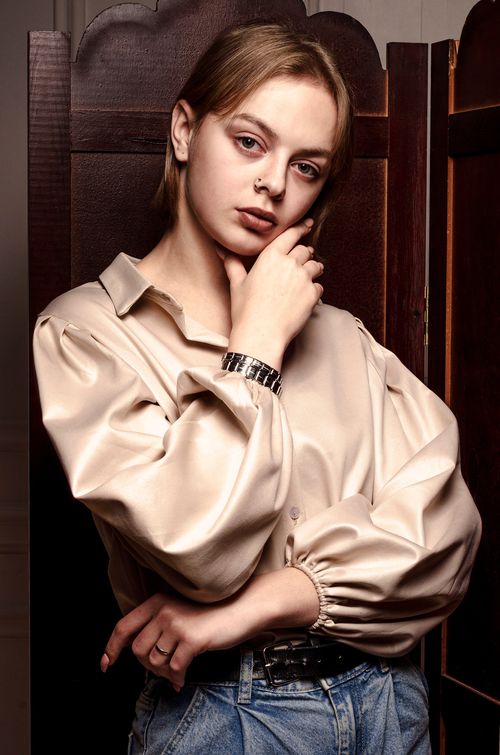 Девушка в задумчивости на фоне ширмы, в бежевой рубашке.