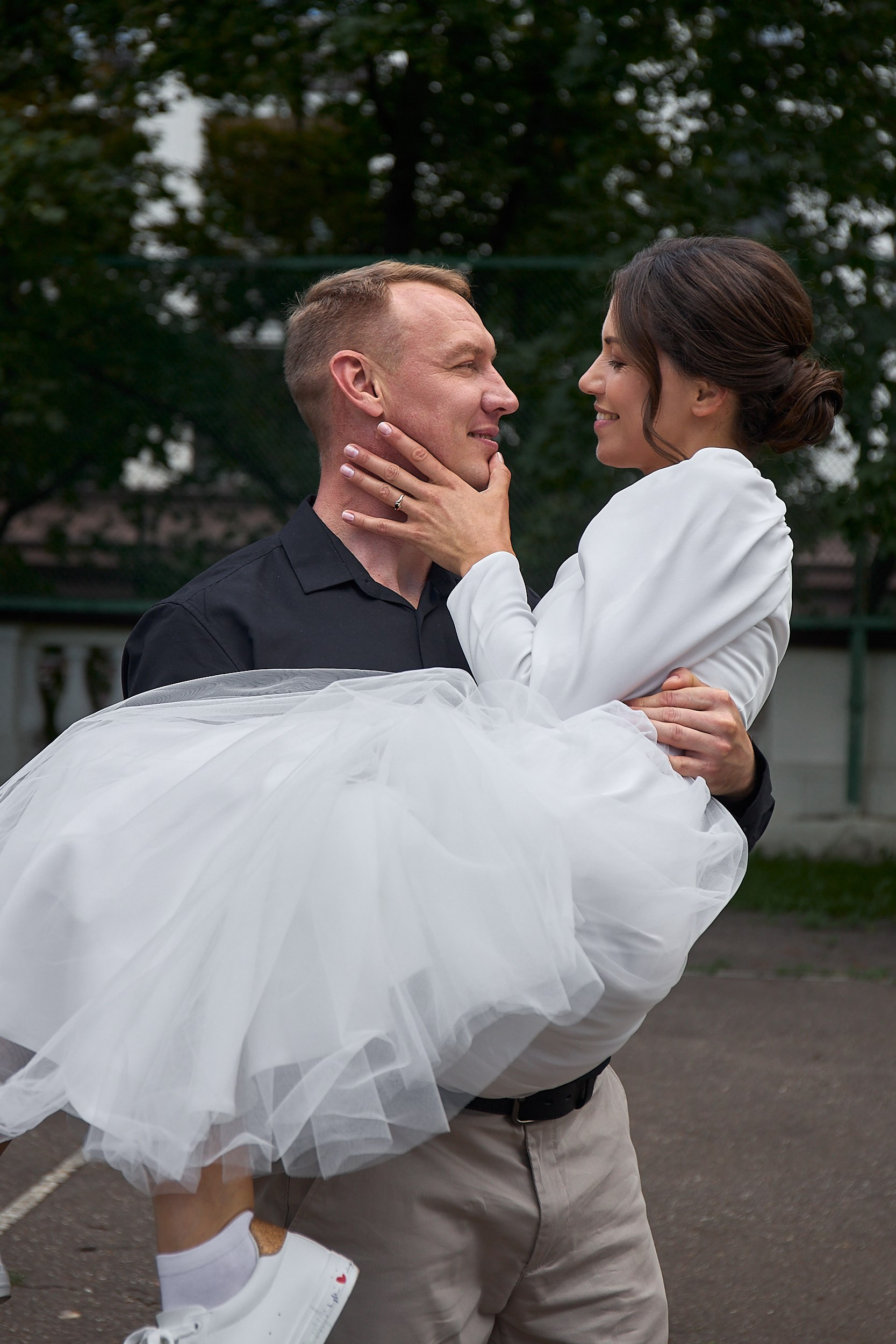 Жених в черной рубашке держит невесту в белом платье на руках.