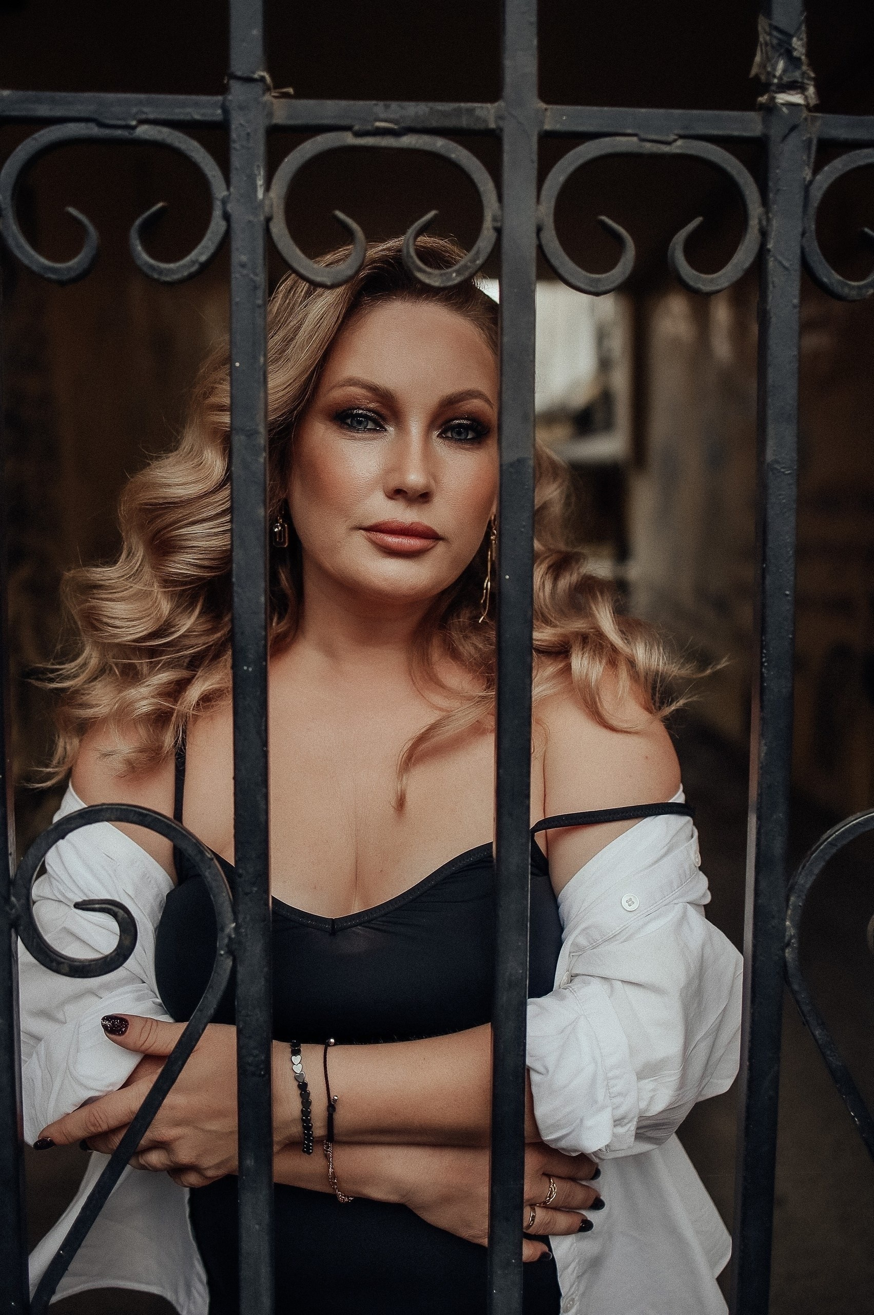 Женщина с волнистыми светлыми волосами стоит за коваными решётками. На ней чёрное платье и белая рубашка.