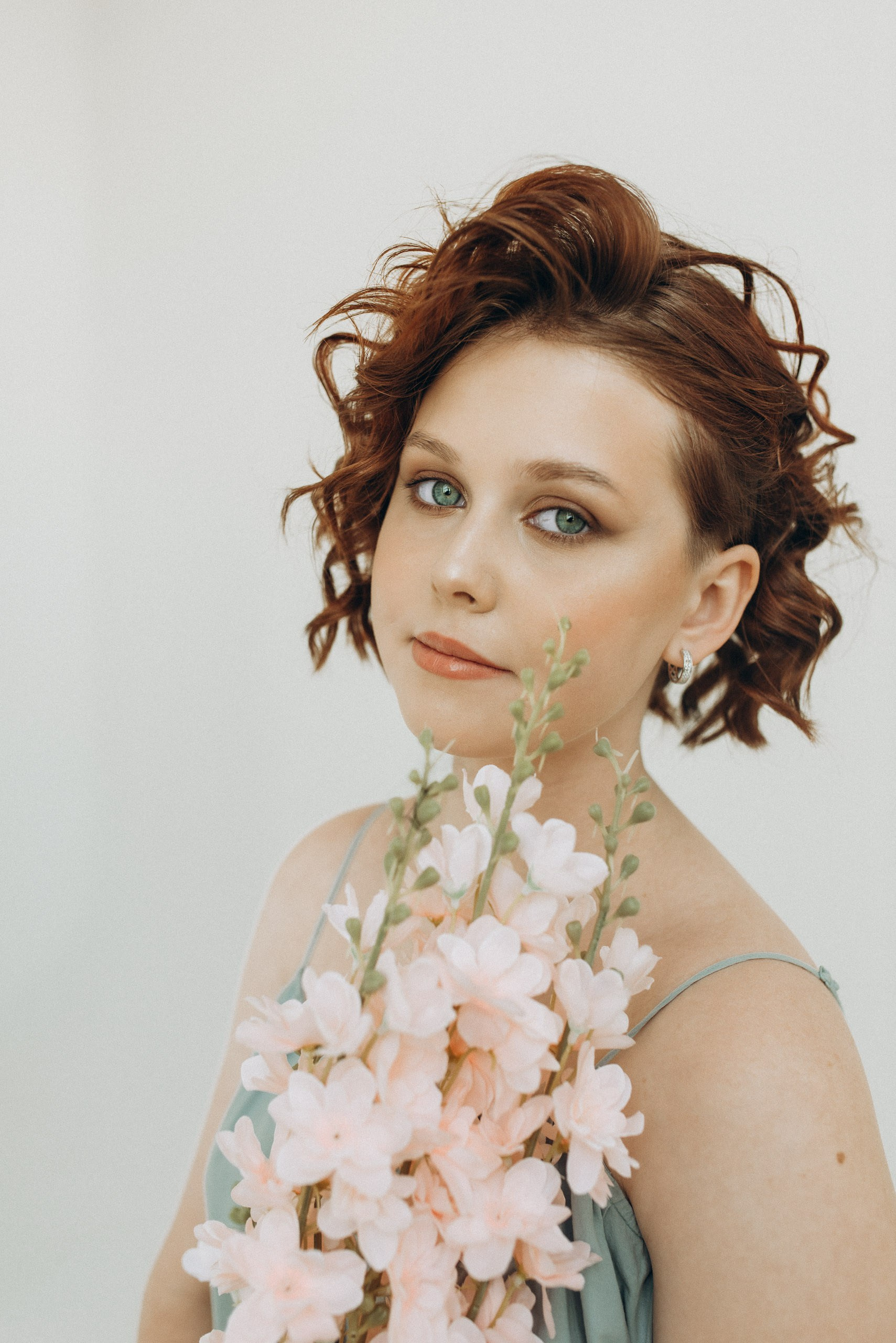 Эстетичная фотосессия для девушки в студии. Рыжие вьющиеся волосы, светлый фон и ветка цветов. Качественное портфолио фотографа