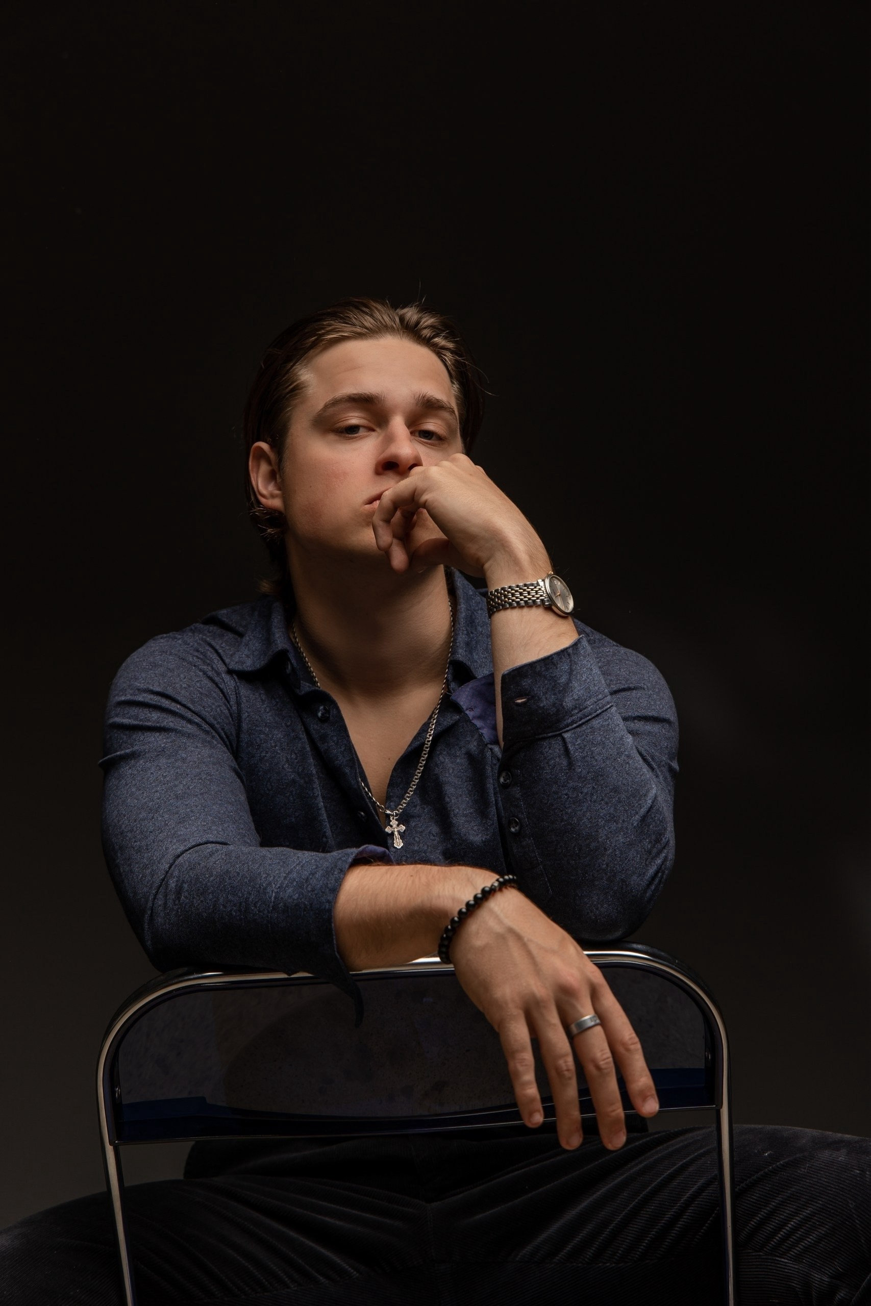 Парень в тёмной рубашке сидит на стуле, задумчиво подперев лицо рукой. Фон тёмный
