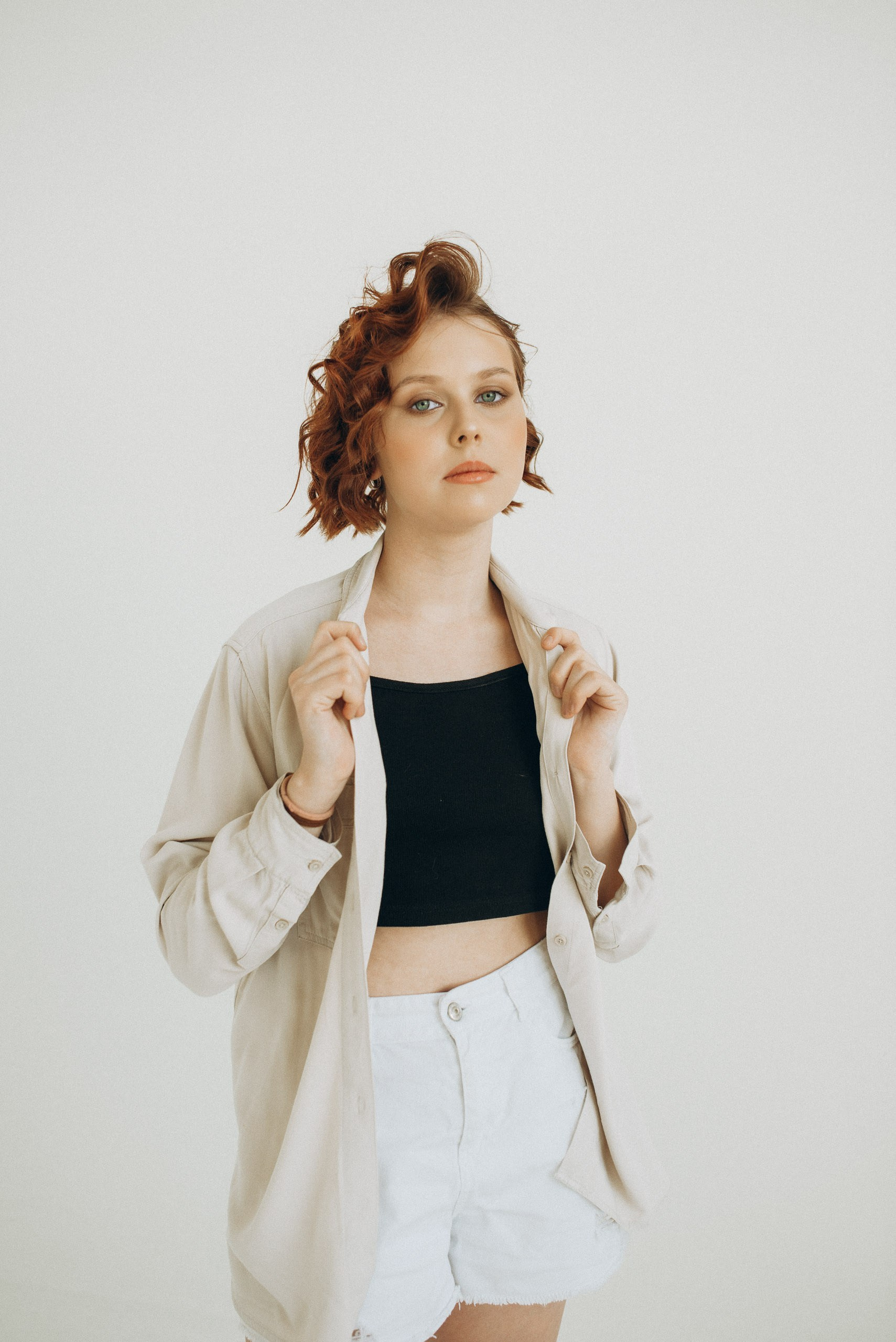 Стильный поясной портрет: девушка в бежевой рубашке, топ-кропе и белых шортах. Руки на воротнике, взгляд в камеру. Молодёжная мода и фотография