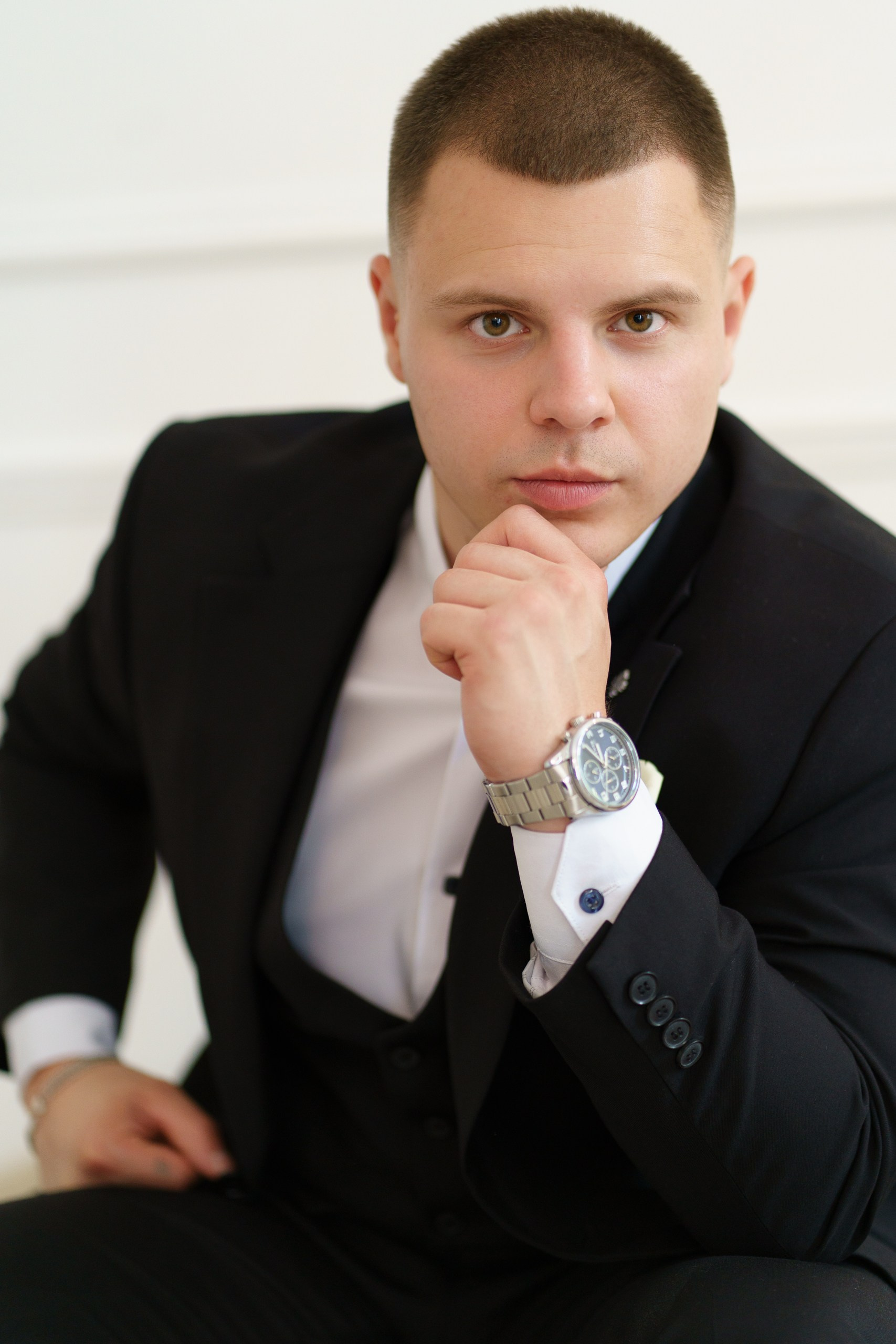 Портрет жениха в классическом стиле: чёрный костюм, белая рубашка, наручные часы. Он сидит, задумчиво опираясь на руку, и смотрит в камеру | свадебная фотосессия Екатеринбург