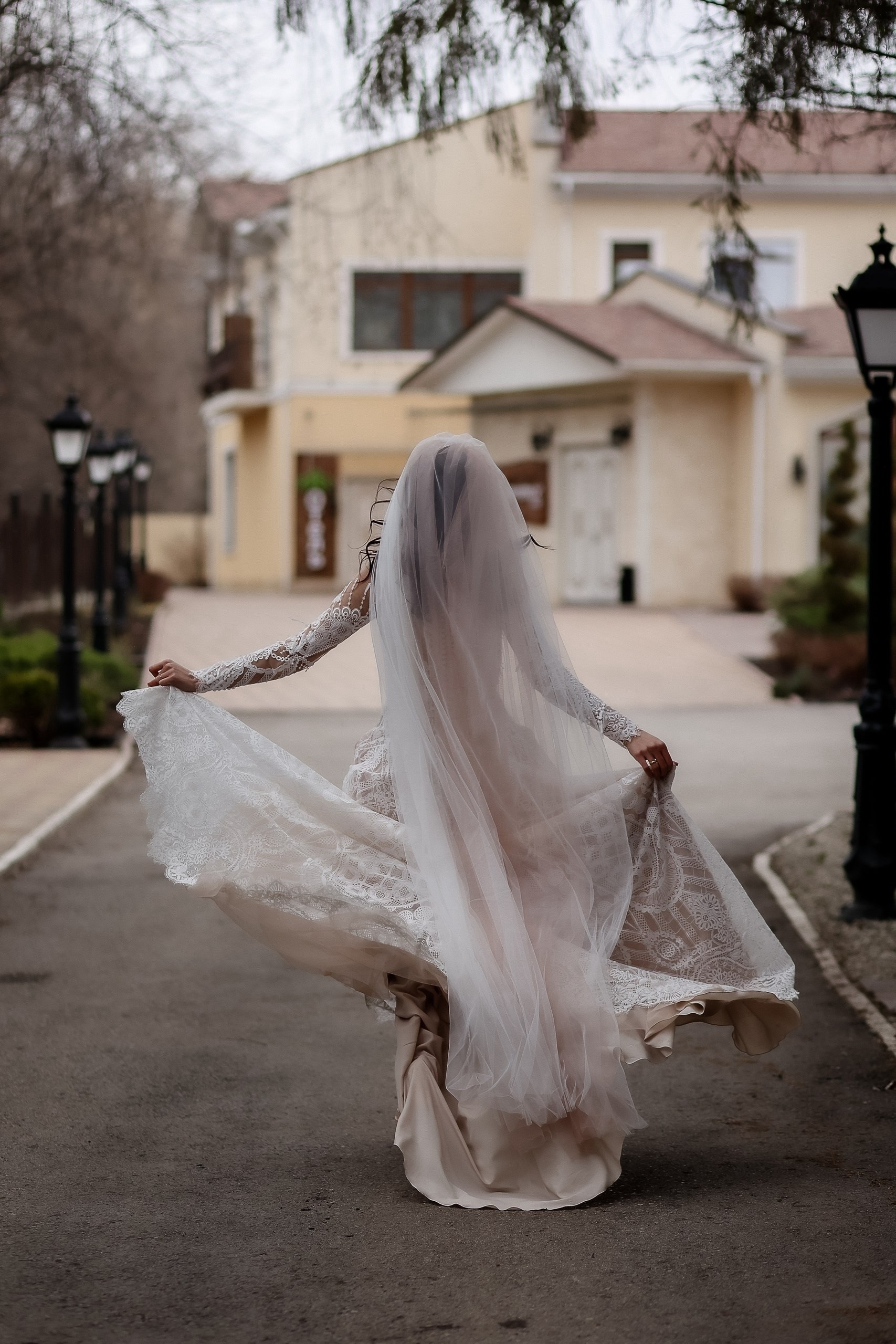 Невеста в пышном свадебном платье с длинной фатой убегает по аллее. На фоне — светлый дом, фонари.