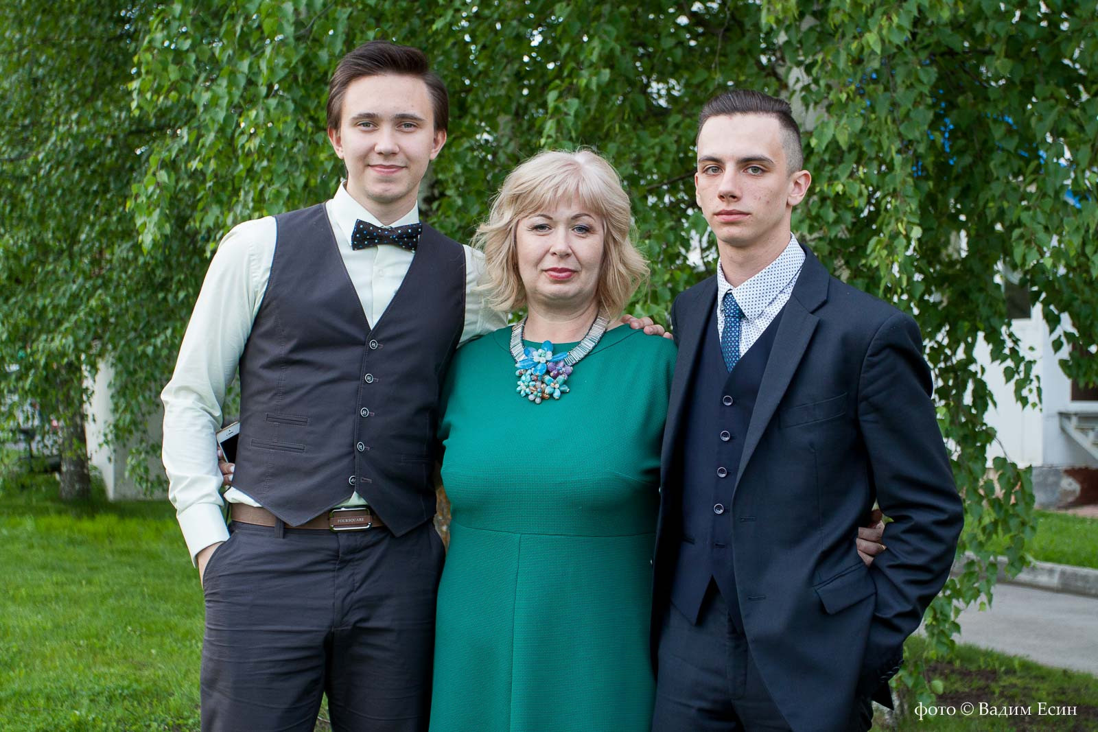 Два выпускника обнимают классного руководителя в зеленом платье на фоне березы.