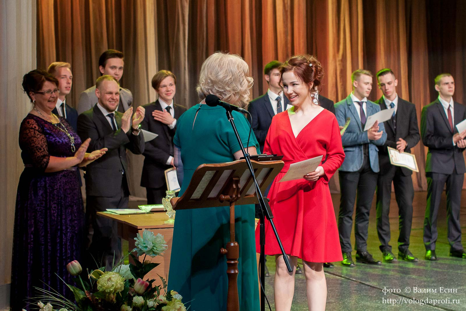 Педагог в зеленом платье торжественно вручает аттестат выпускнице 11 класса в красном платье.