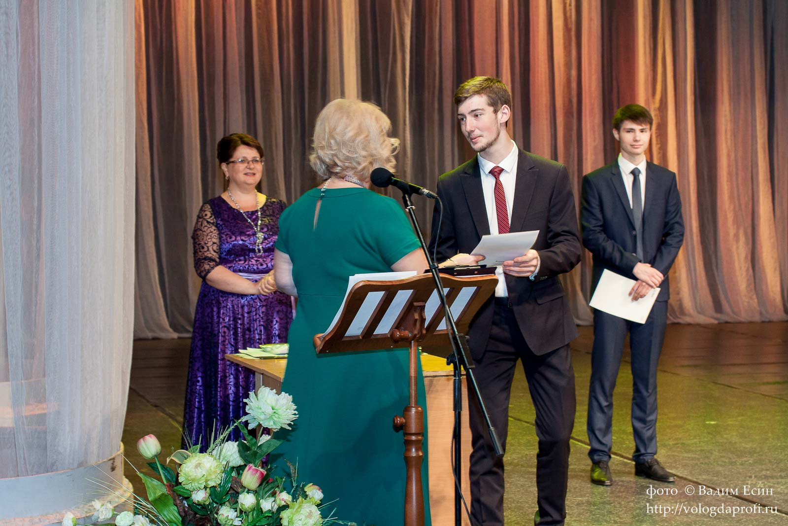 Педагог в зеленом платье торжественно вручает аттестат выпускнику 11 класса в темном костюме и красном галстуке.