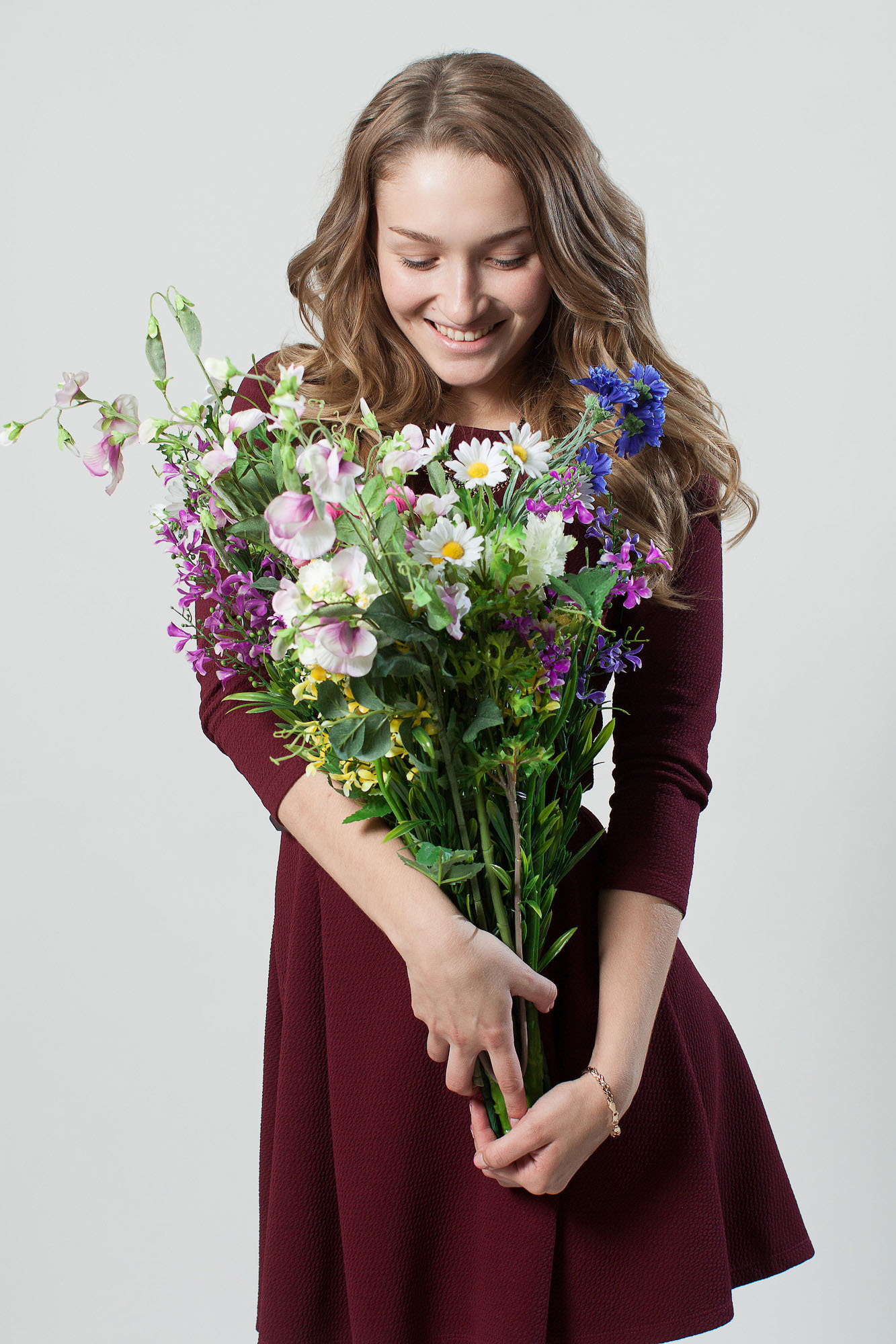 Поколенный студийный портрет девушки с с букетом цветов в руках в бордовом платье на светло-сером фоне