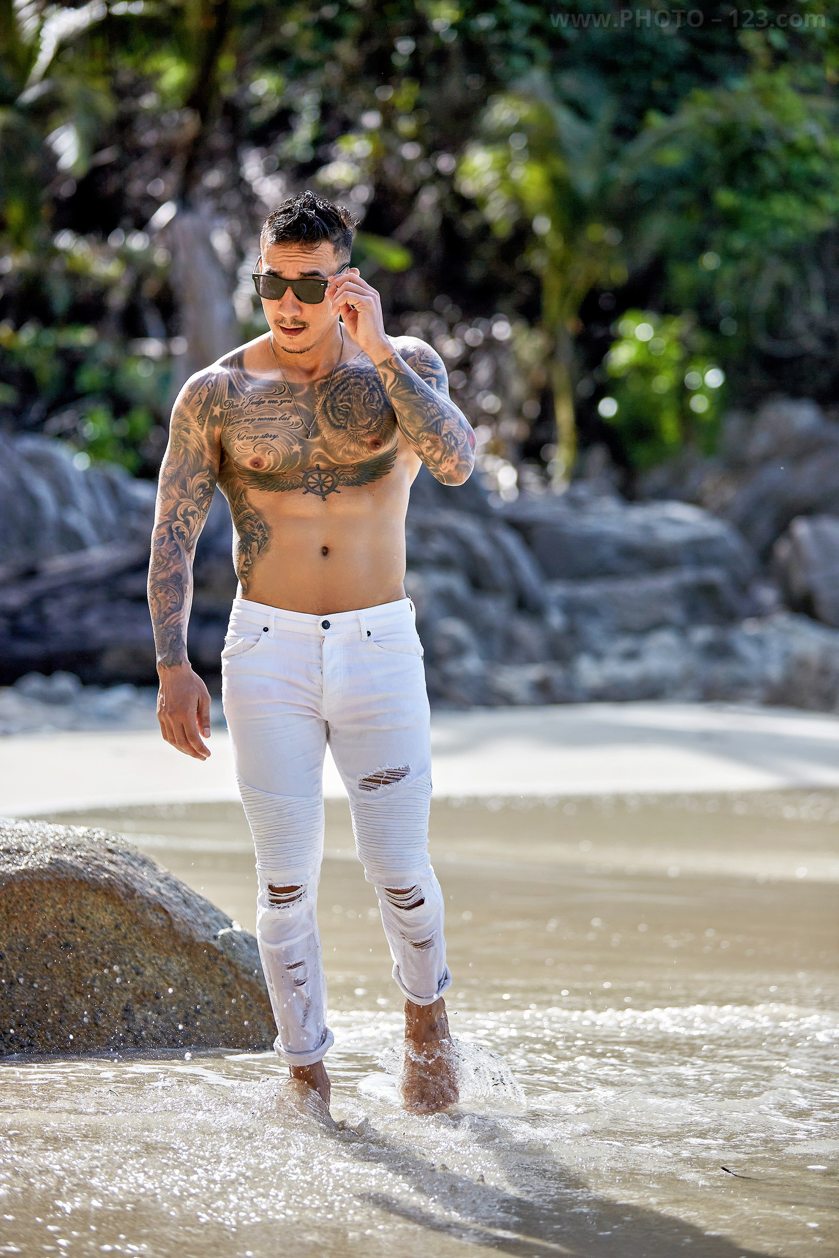 Shirtless athletic male model adjusting sunglasses while walking in ocean waves on a Nha Trang beach