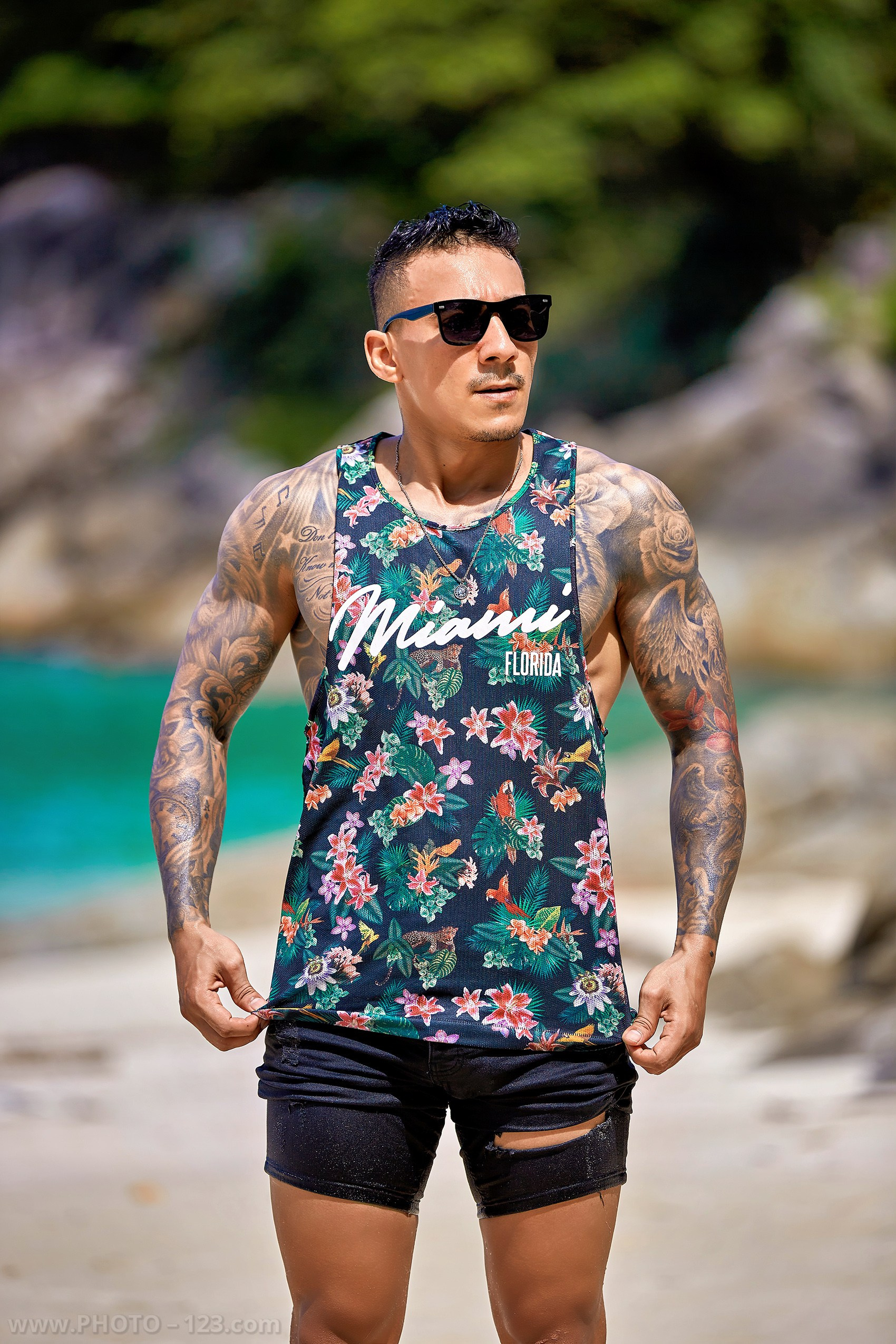Tattooed man in floral tank top and sunglasses posing on a Nha Trang beach