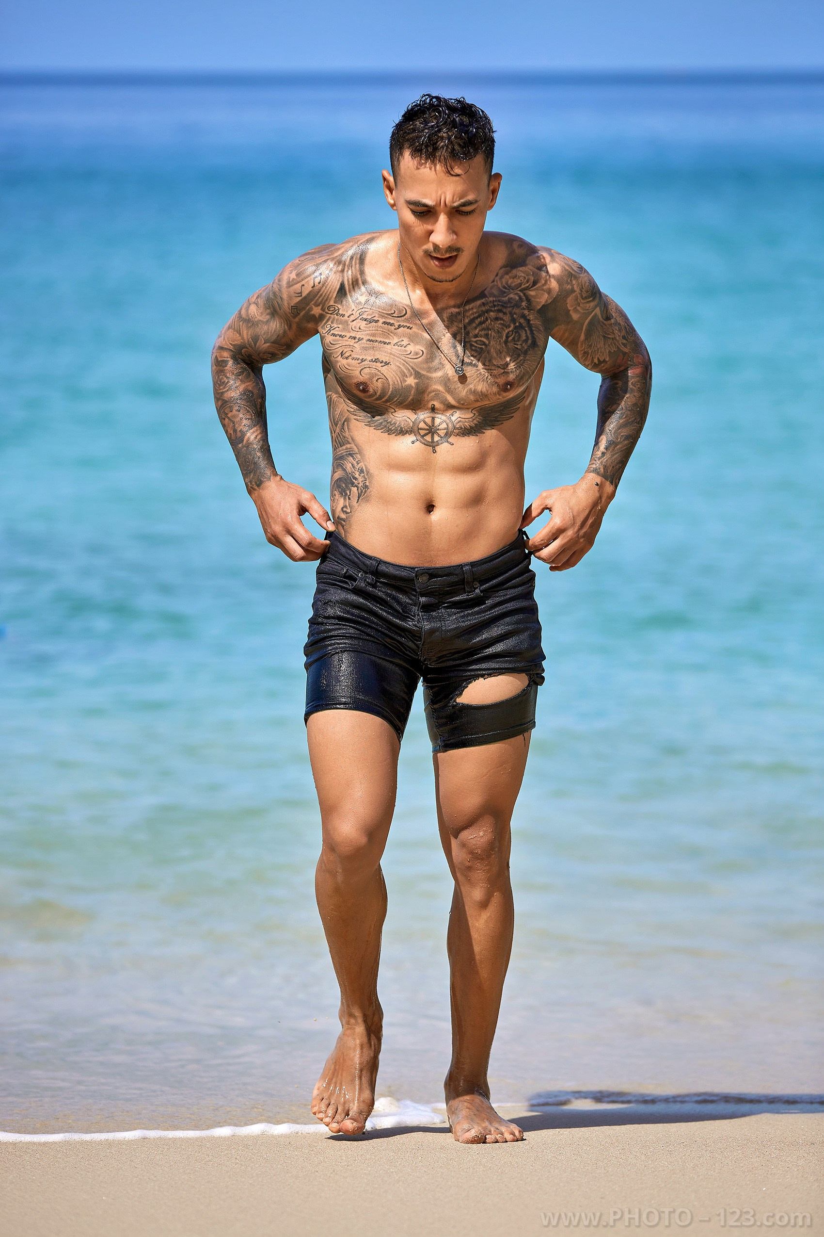Tattooed fitness model adjusting his shorts while walking along the shoreline in Nha Trang