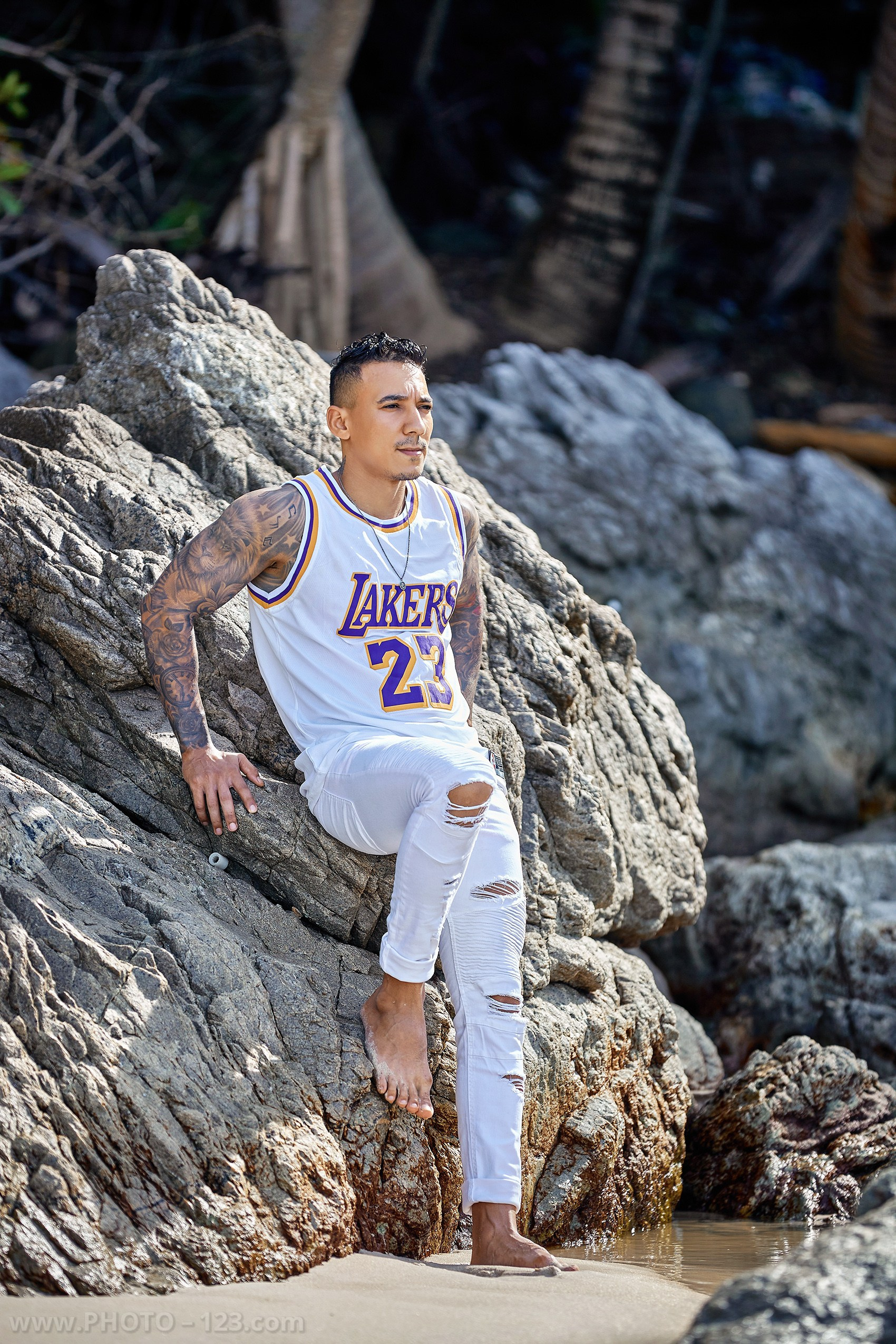 Athletic tattooed man in a white basketball jersey posing on rocky beach in Nha Trang by professional photographer