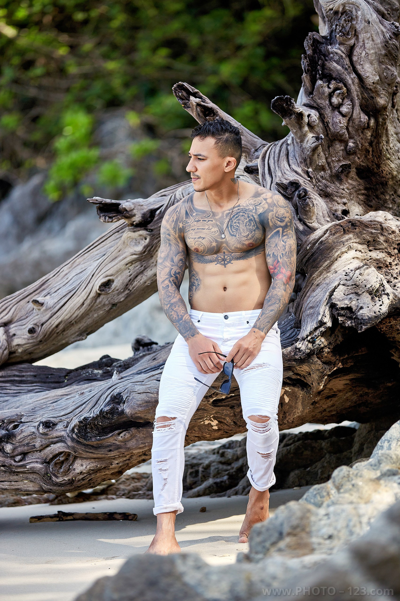 Muscular tattooed man standing next to large coastal driftwood in Nha Trang by professional photographer