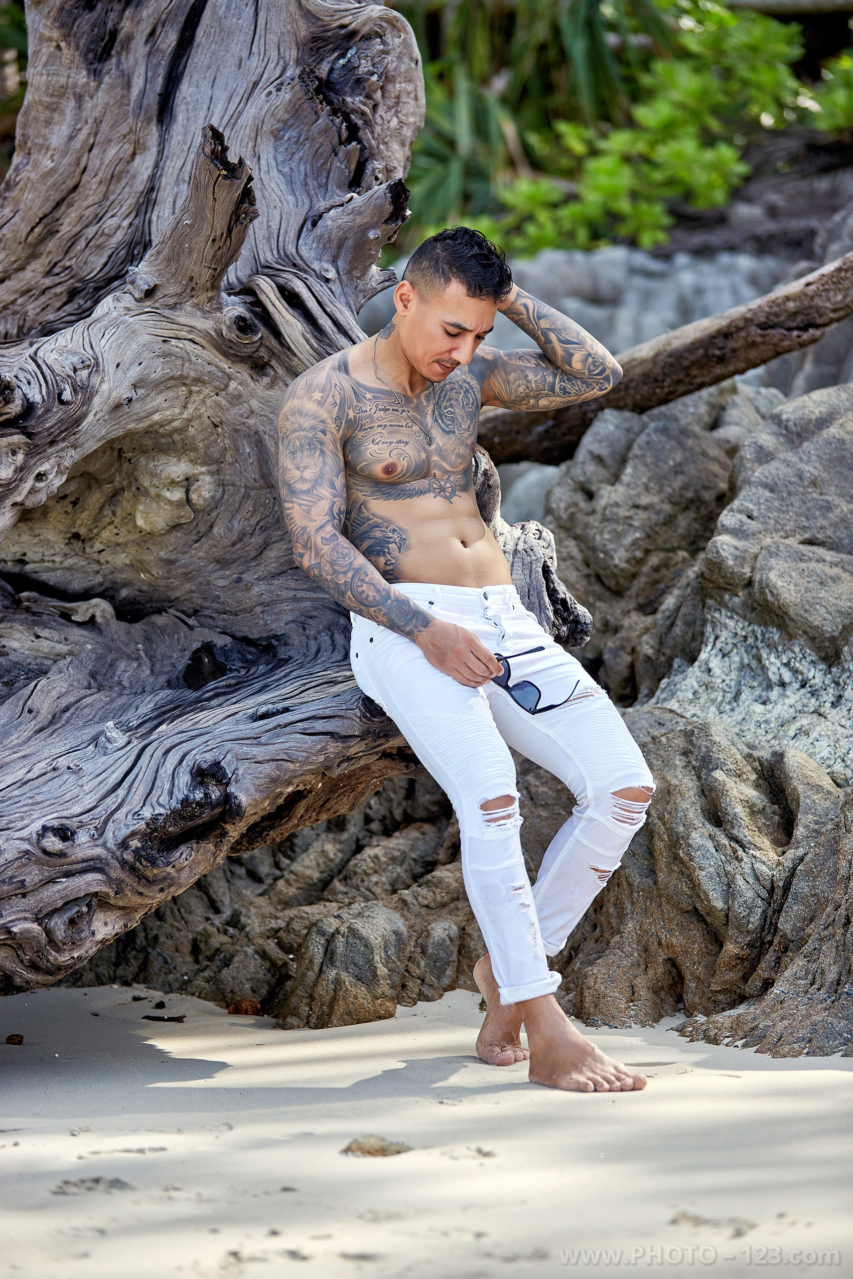 Tattooed man leaning against large weathered driftwood on a beach during a Nha Trang portrait session