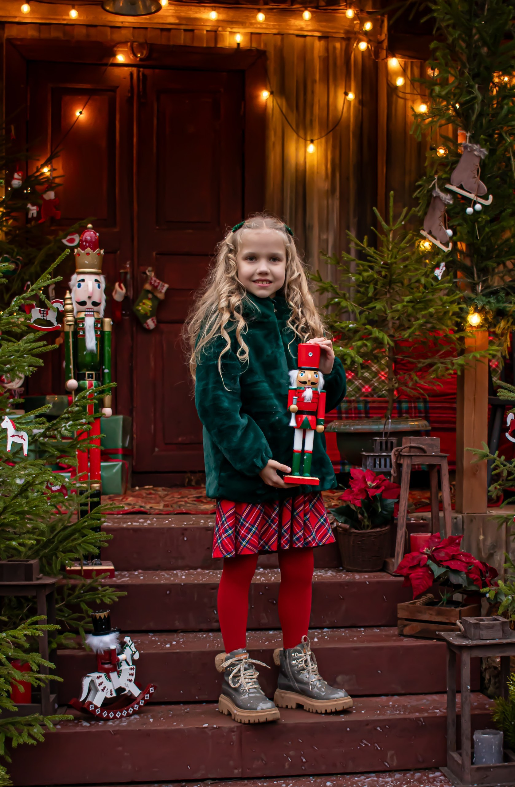 Девочка в зелёной шубке улыбается на новогодней локации среди ёлок, фотограф Карасёва Елена