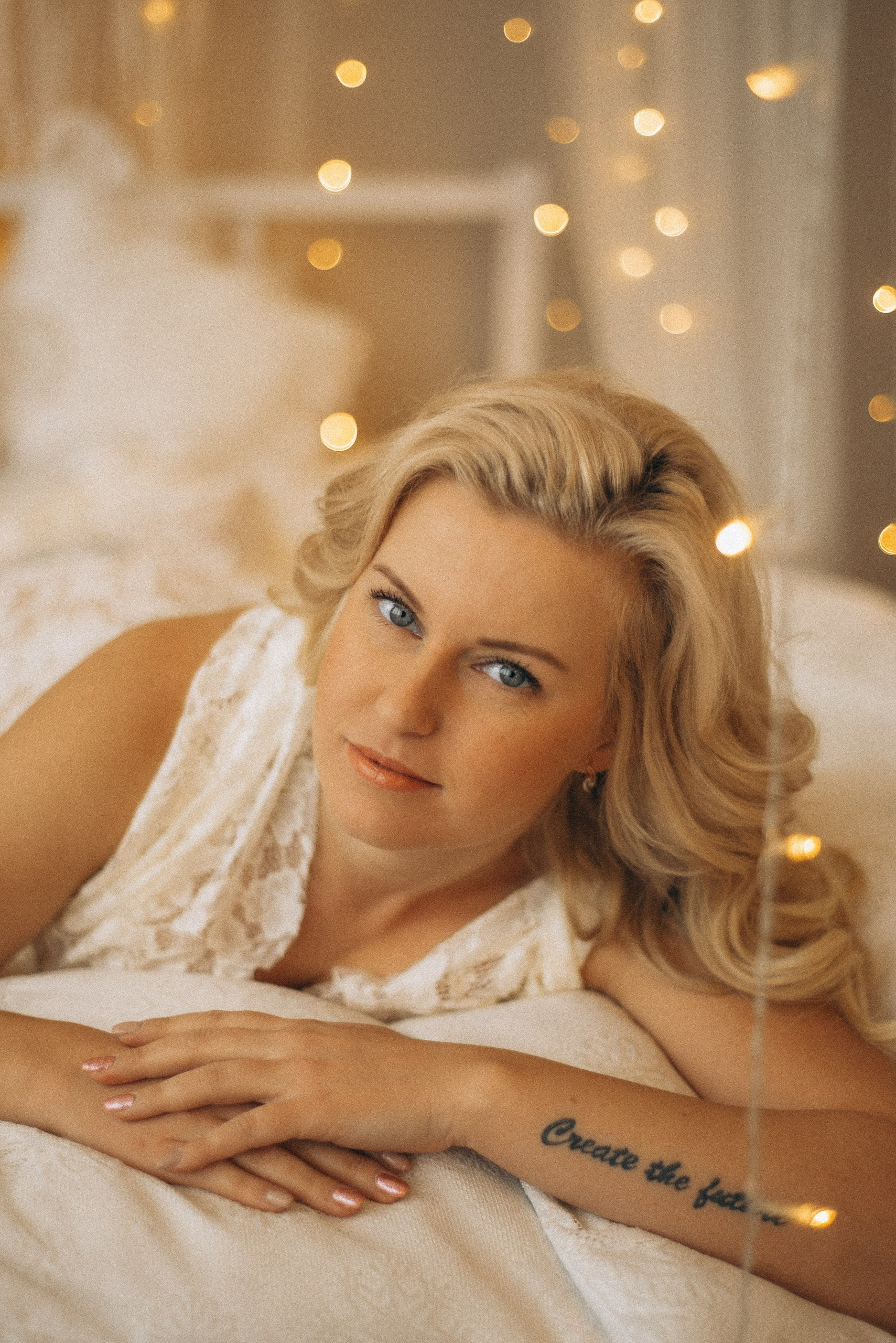 Крупный портрет Натали Каролин на кровати. Акцент на татуировке на руке, текстуре белого кружевного платья и светлом маникюре. Мягкий свет гирлянд на фоне бохо-студии. Женская фотосессия в Новосибирске