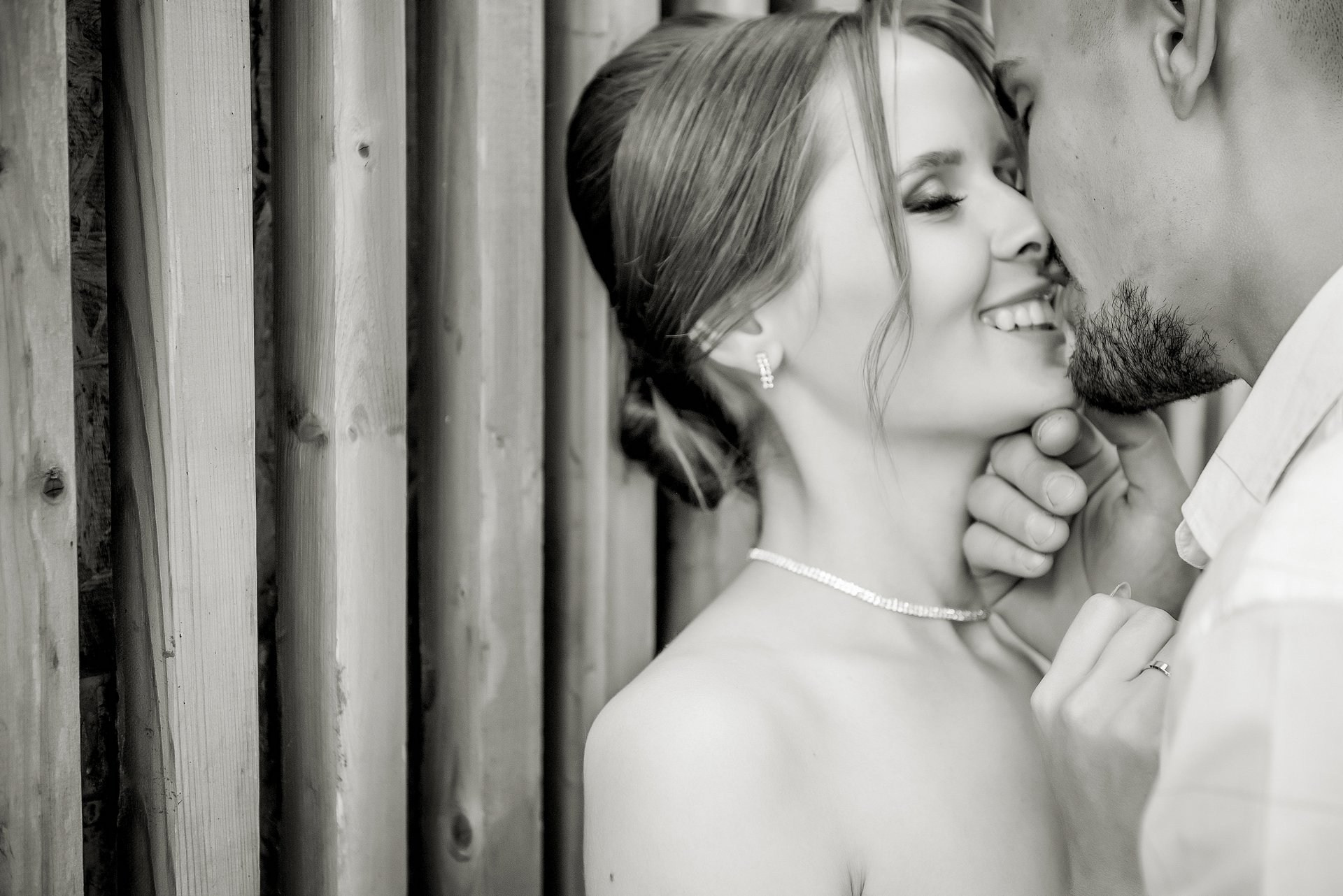 Черно-белое фото мужчины и женщины, стоящих рядом и улыбающихся друг другу, женщина держит руку у лица мужчины. На заднем плане деревянный забор.