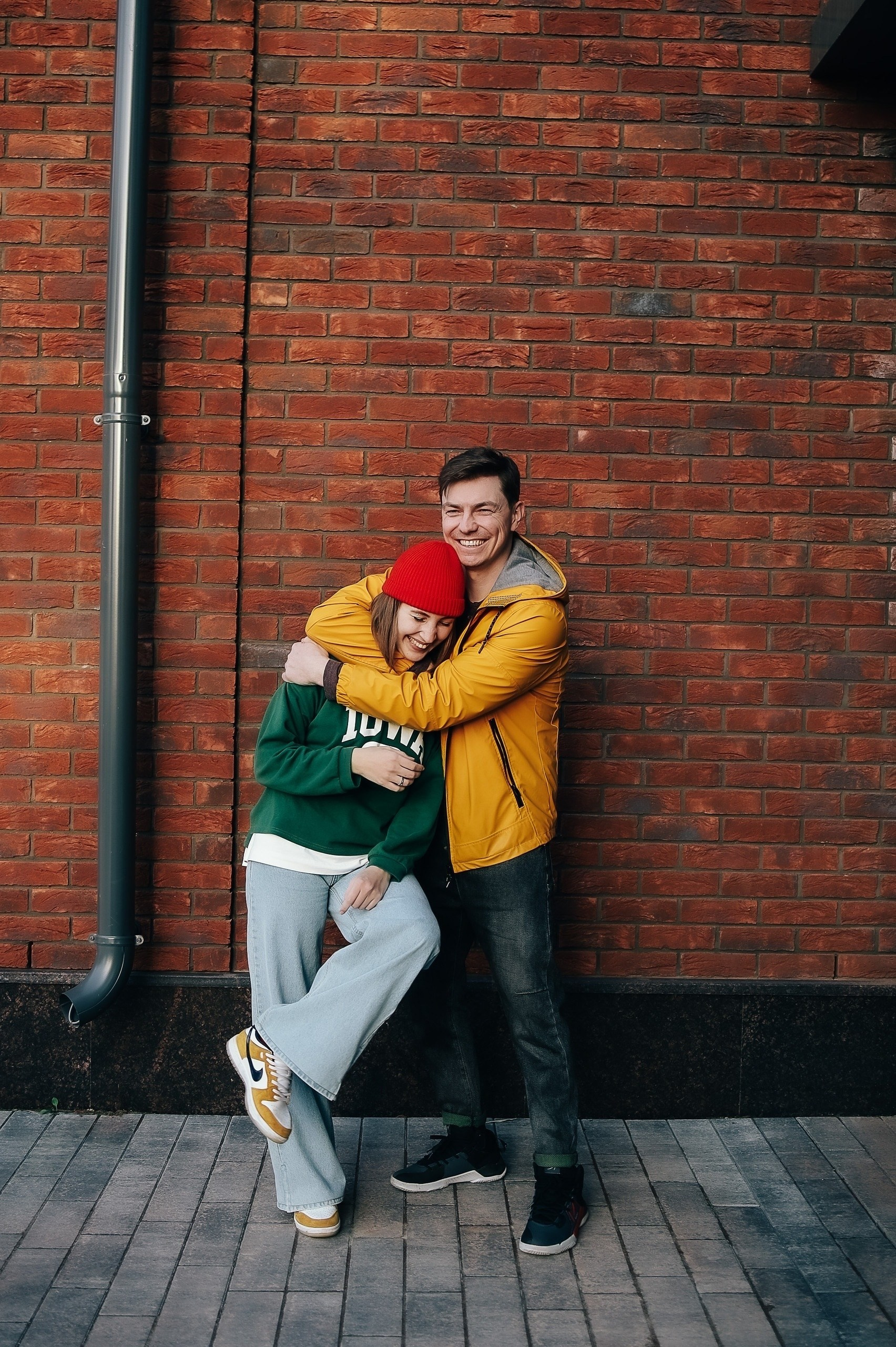 Парень в жёлтой куртке обнимает девушку в красной шапке и зелёной толстовке. Стоят у кирпичной стены, улыбаются.