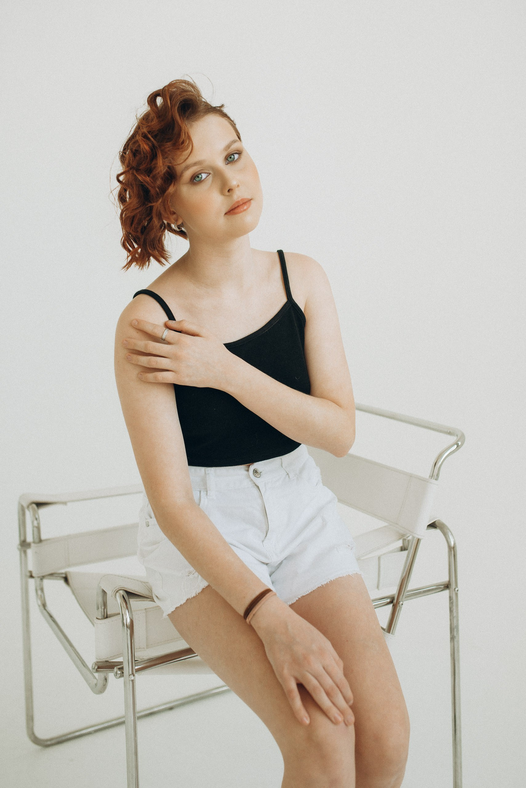 Девушка с рыжими локонами сидит на дизайнерском белом стуле. Чёрный топ, белые шорты и взгляд в камеру. Стильный студийный портрет в Новосибирске