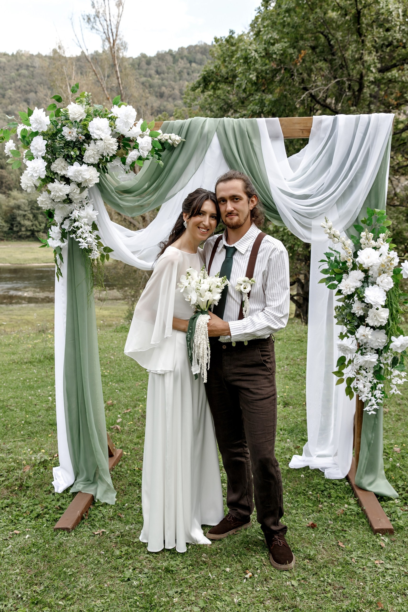 Молодожёны у свадебной арки на природе — он в подтяжках и галстуке, она в лёгком белом платье с букетом. Атмосфера романтичная, вокруг зелень и река.