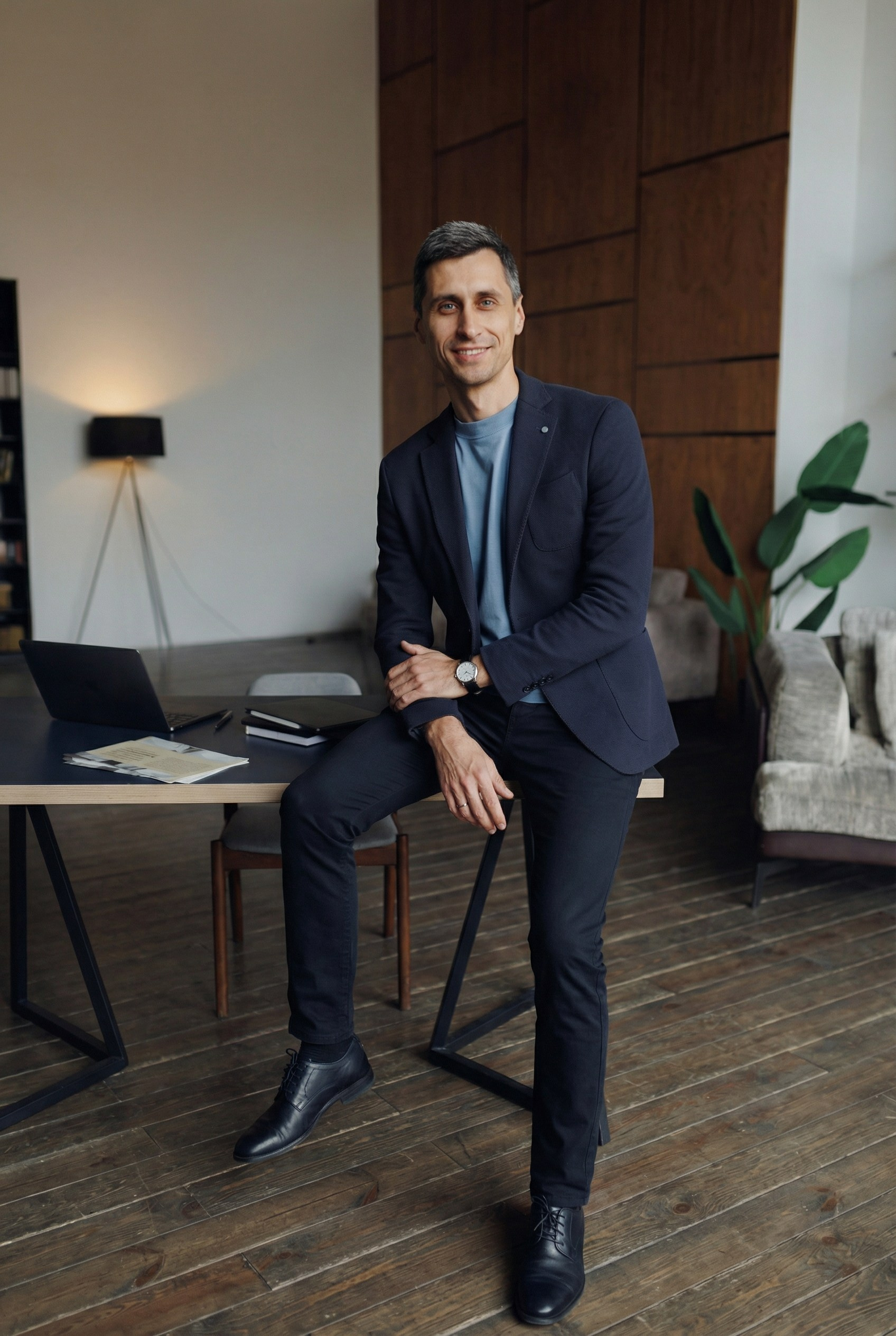 Мужчина во весь рост сидит полусидя на столе в интерьере офиса