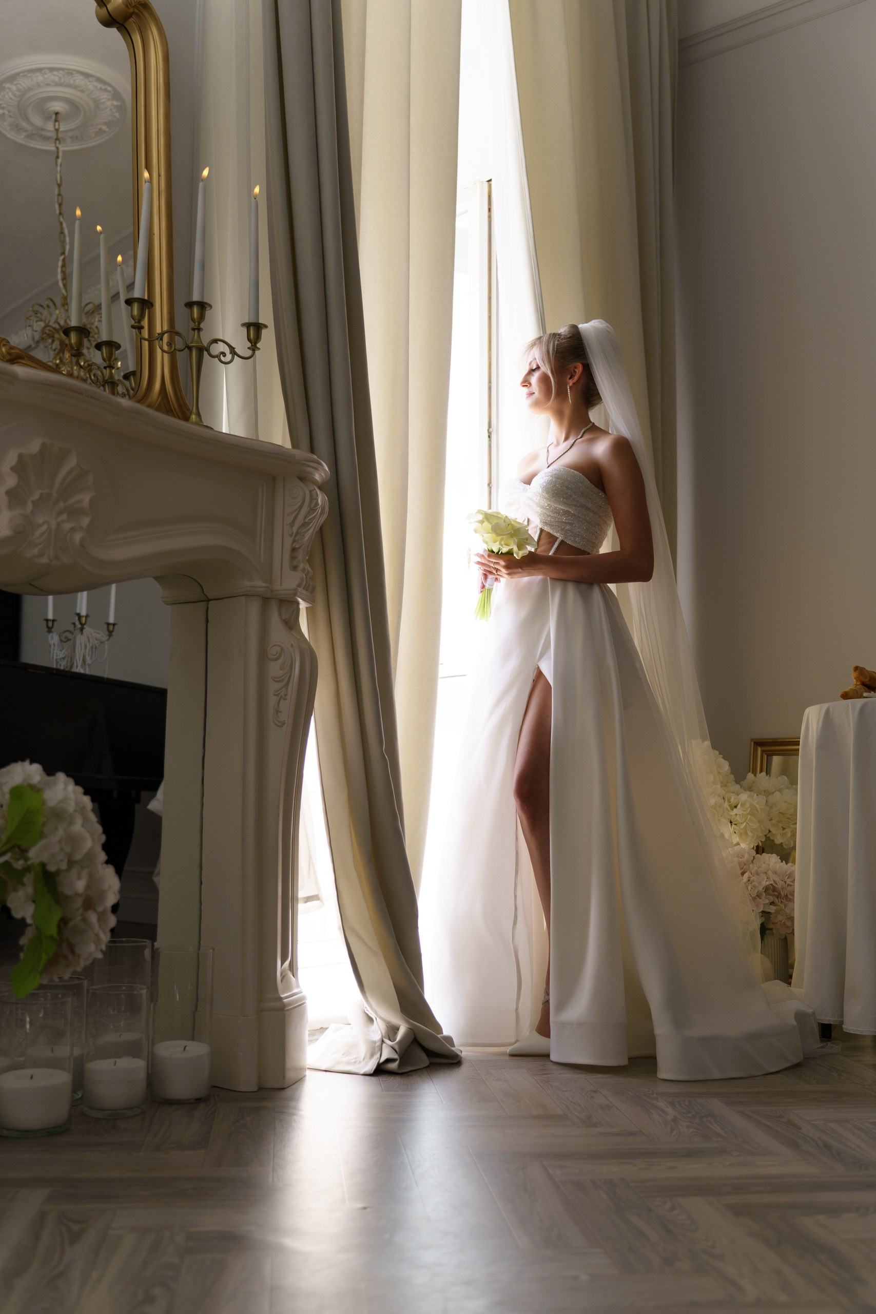 Свадебная фотосессия: невеста в изысканном платье с разрезом грациозно позирует у окна. Мягкий дневной свет подчёркивает её образ. На переднем плане — свечи в стеклянных подсвечниках и букет гортензий | креативный свадебный фотограф Екатеринбург
