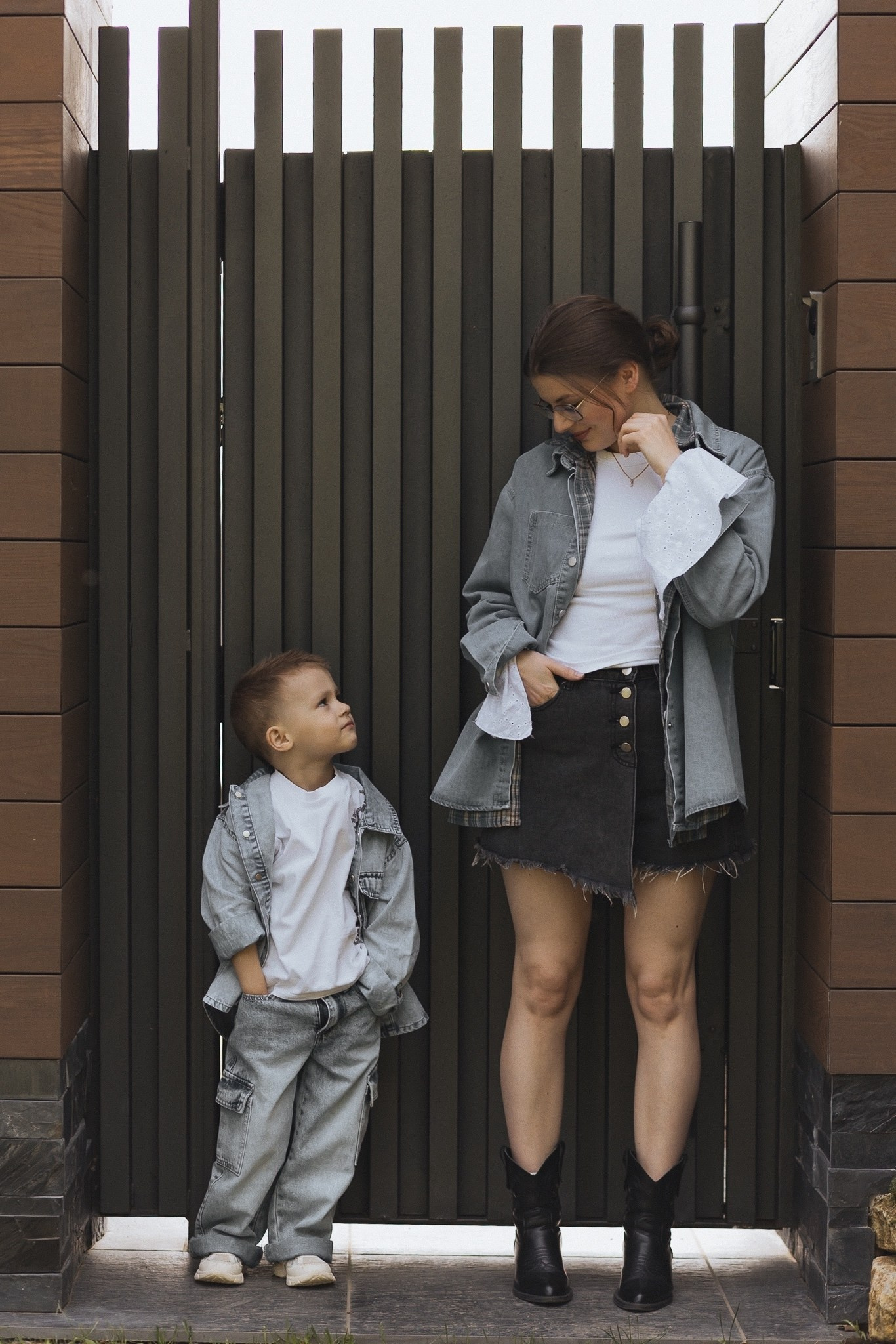 Мама и сын стоят у стильных тёмных ворот. Оба в джинсовой одежде, выглядят модно.