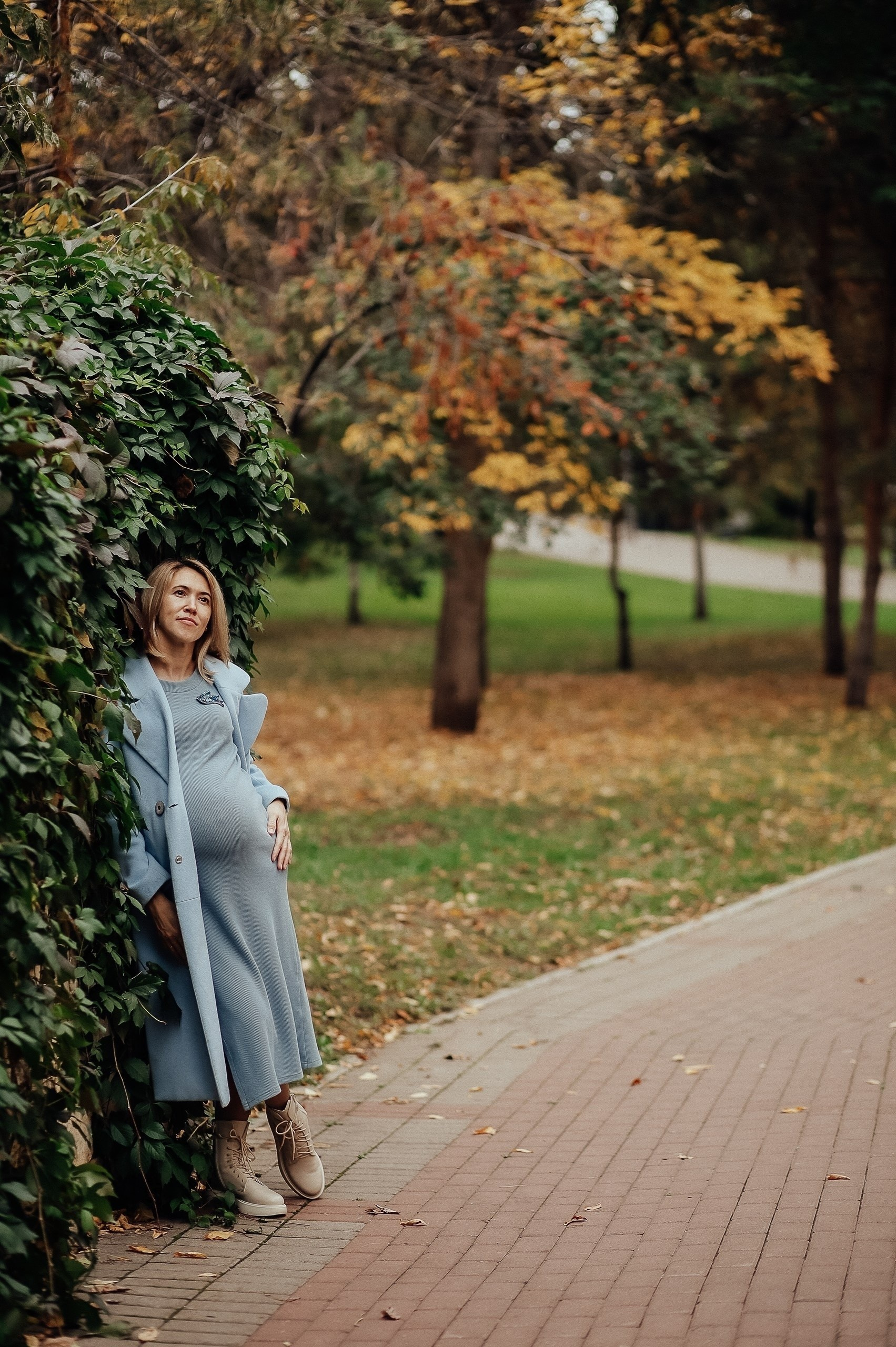 Беременная женщина в голубом платье стоит в парке у живой изгороди. Вокруг осенние деревья.