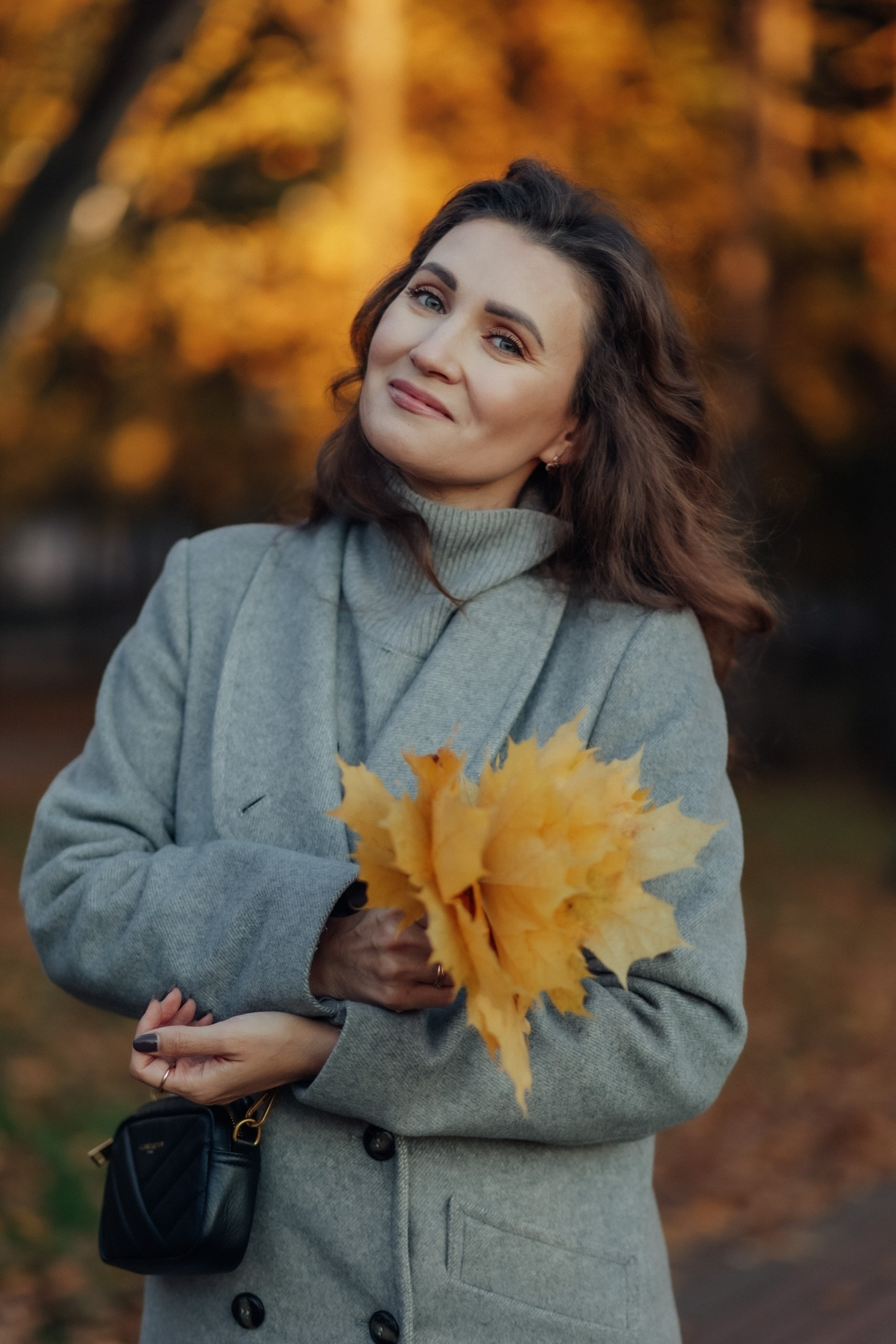 Женщина в сером пальто держит букет жёлтых кленовых листьев. Стоит в осеннем парке, выглядит задумчивой и счастливой.