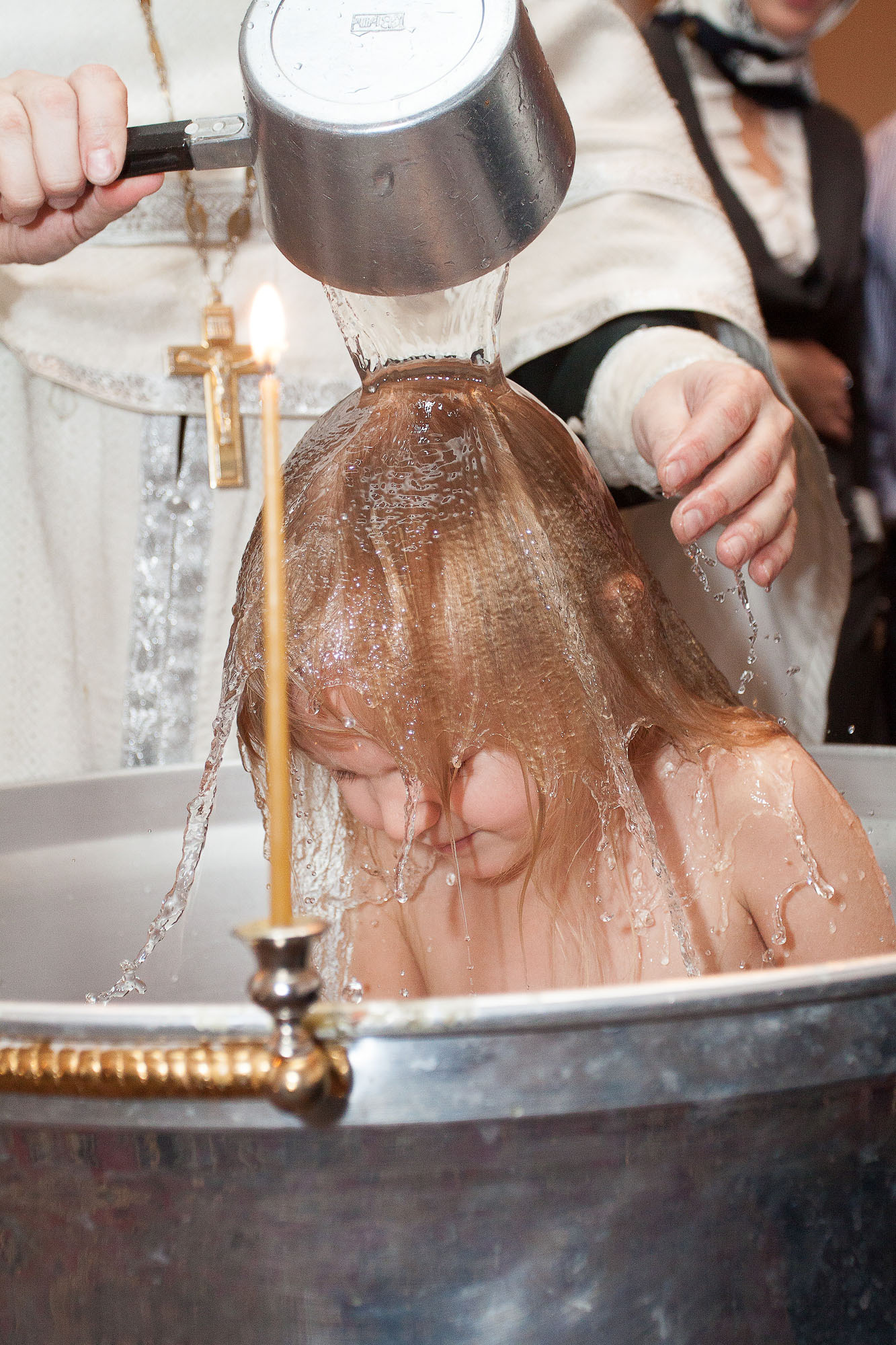 Священник выливает воду из серебристого ковшика на голову девочке, которая стоит в чане.