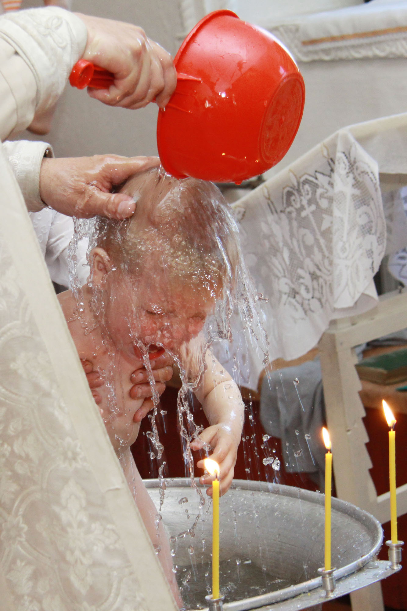 Священник выливает воду из красного ковшика на голову ребенка, который стоит в чане.