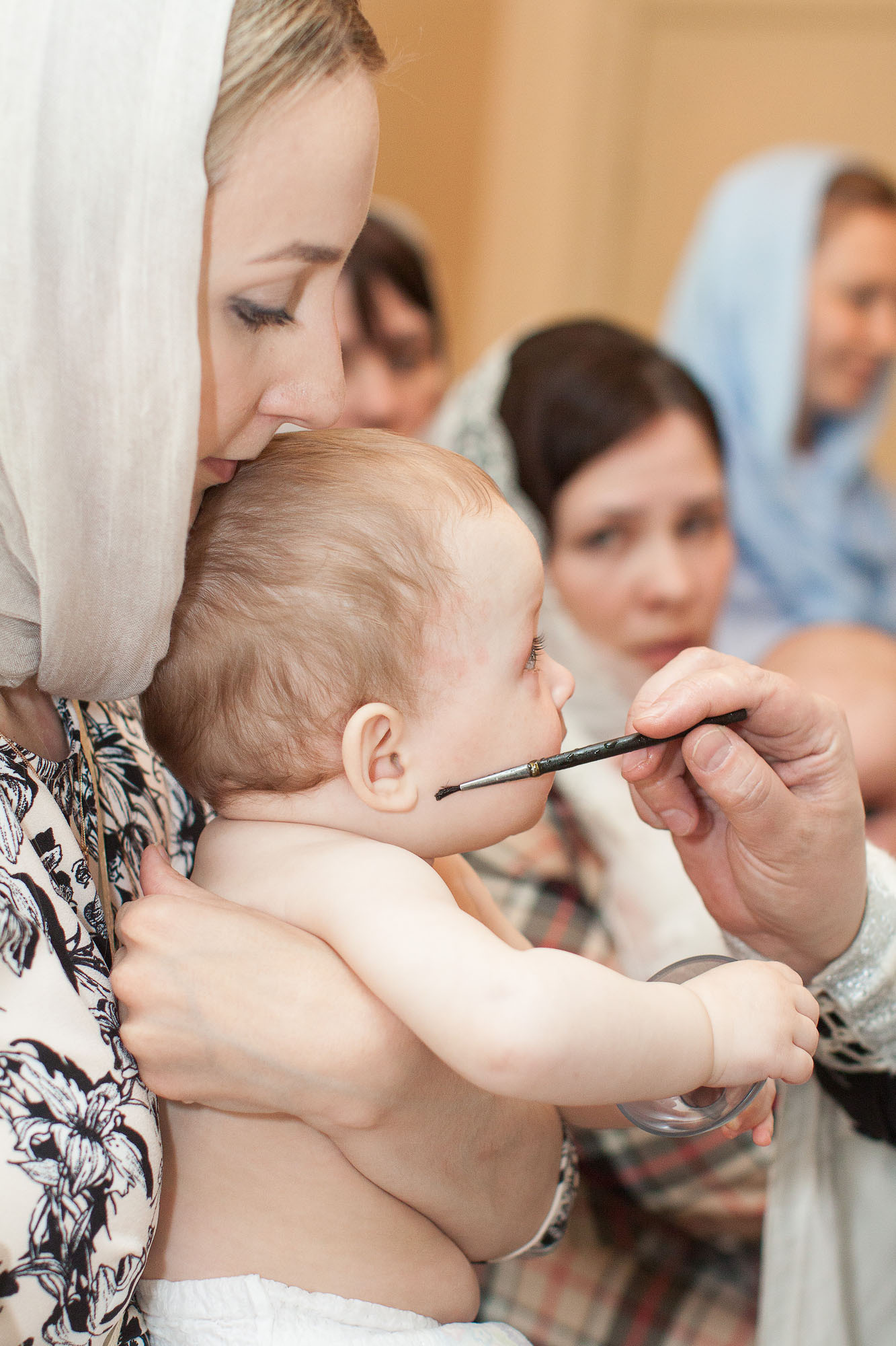 Рука священника с кисточкой касается правой щеки малыша, который сидит на руках у женщины в платке.